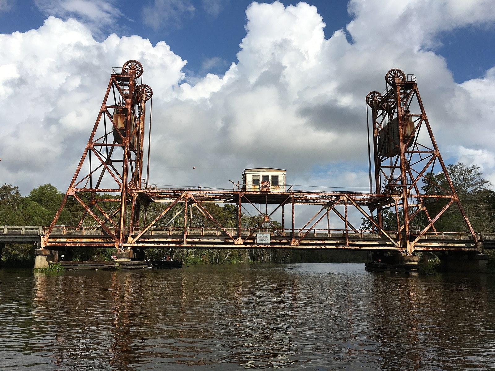 Rusty, industrial drawbridge spans a calm river under a partly cloudy sky.