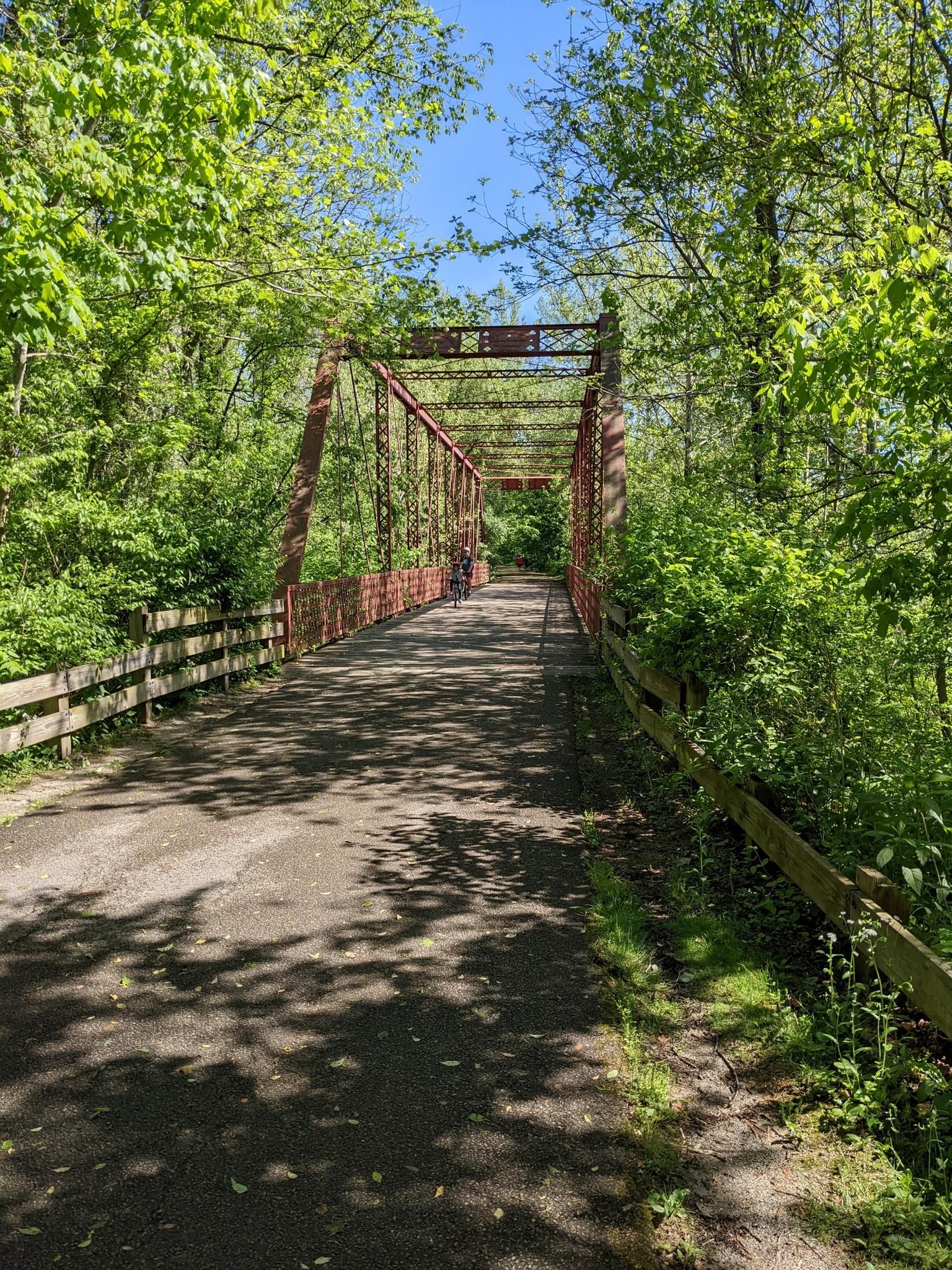Paved path leading to an old red metal bridge, surrounded by green trees. - Limestone Greenway
