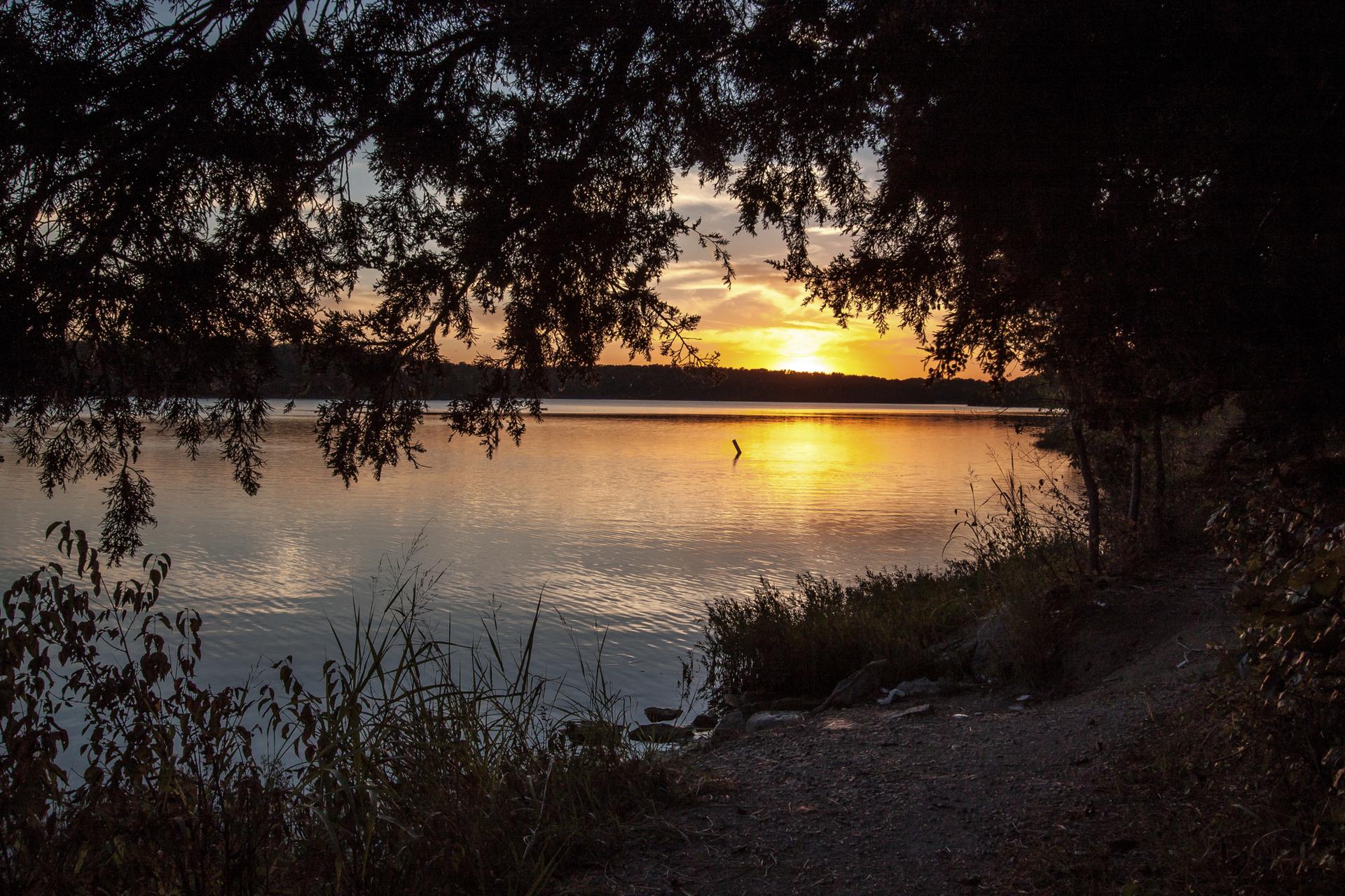 Sunset over a calm lake, framed by dark trees and glowing water.