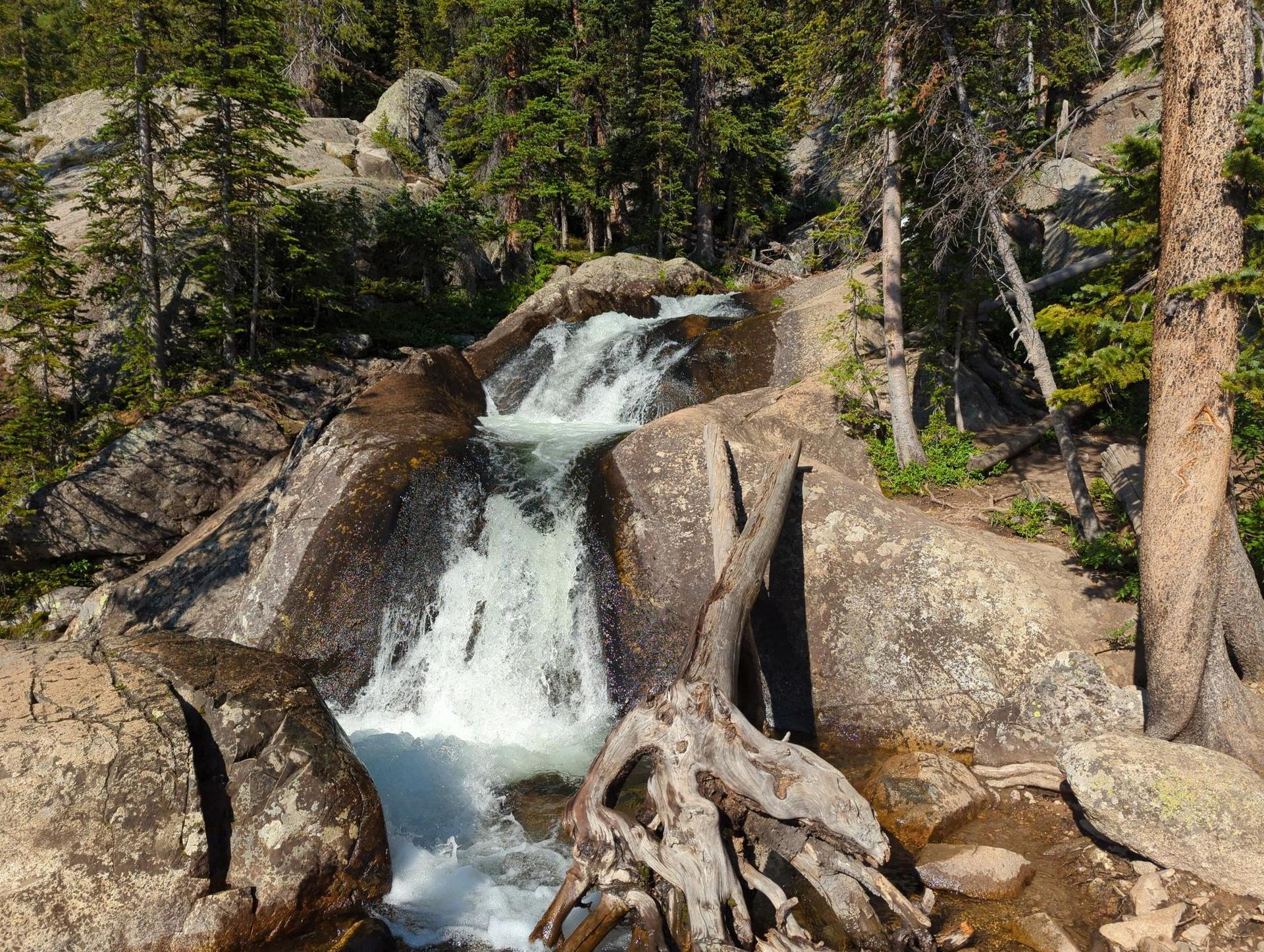 Multi-tiered waterfall cascades over large rocks in a sunny, green forest.