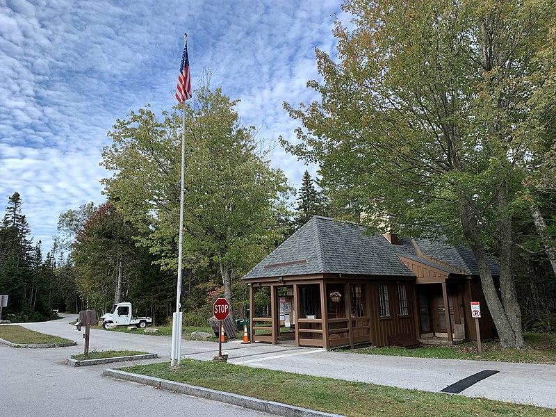 Wooden building, American flag on pole, and paved road surrounded by trees.