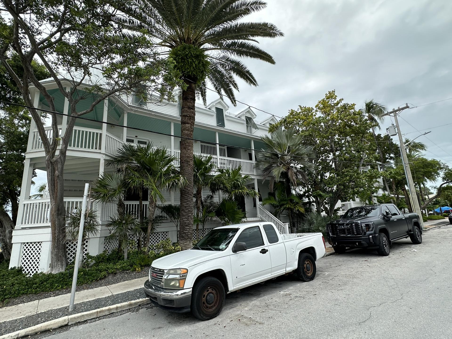 White house with balconies, palm trees, and parked trucks on a street.