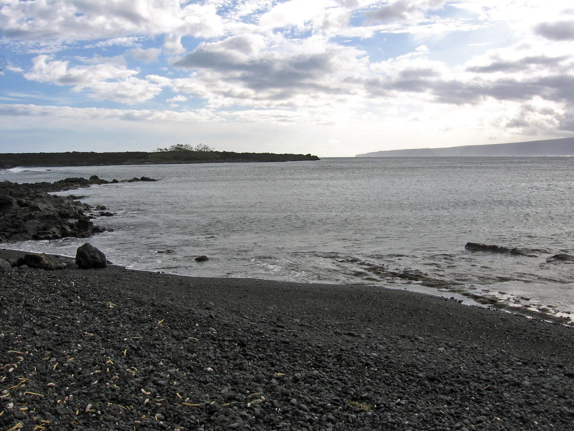 Dark volcanic beach meets calm ocean under a bright, cloudy sky.
