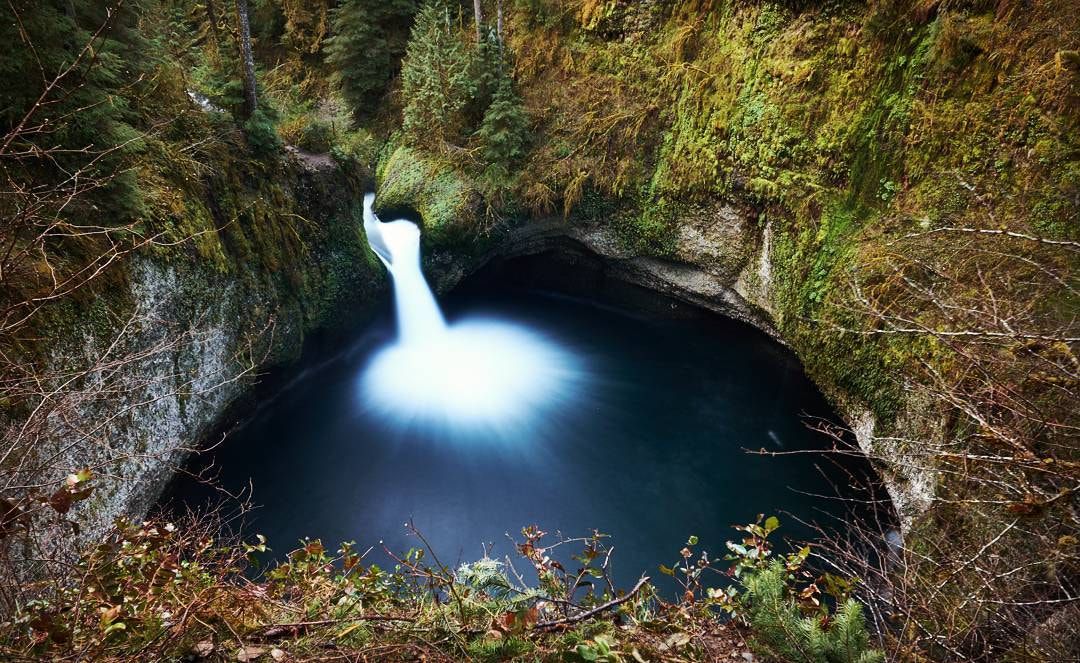 Waterfall plunges into a dark, glowing pool within a mossy, rocky grotto. - Punchbowl Falls – Punchbowl Falls loop from Dee