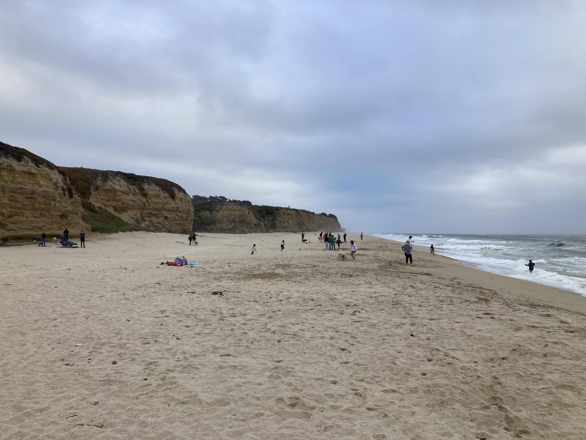 Crowded sandy beach with people, crashing waves, and tall eroded cliffs under cloudy sky.