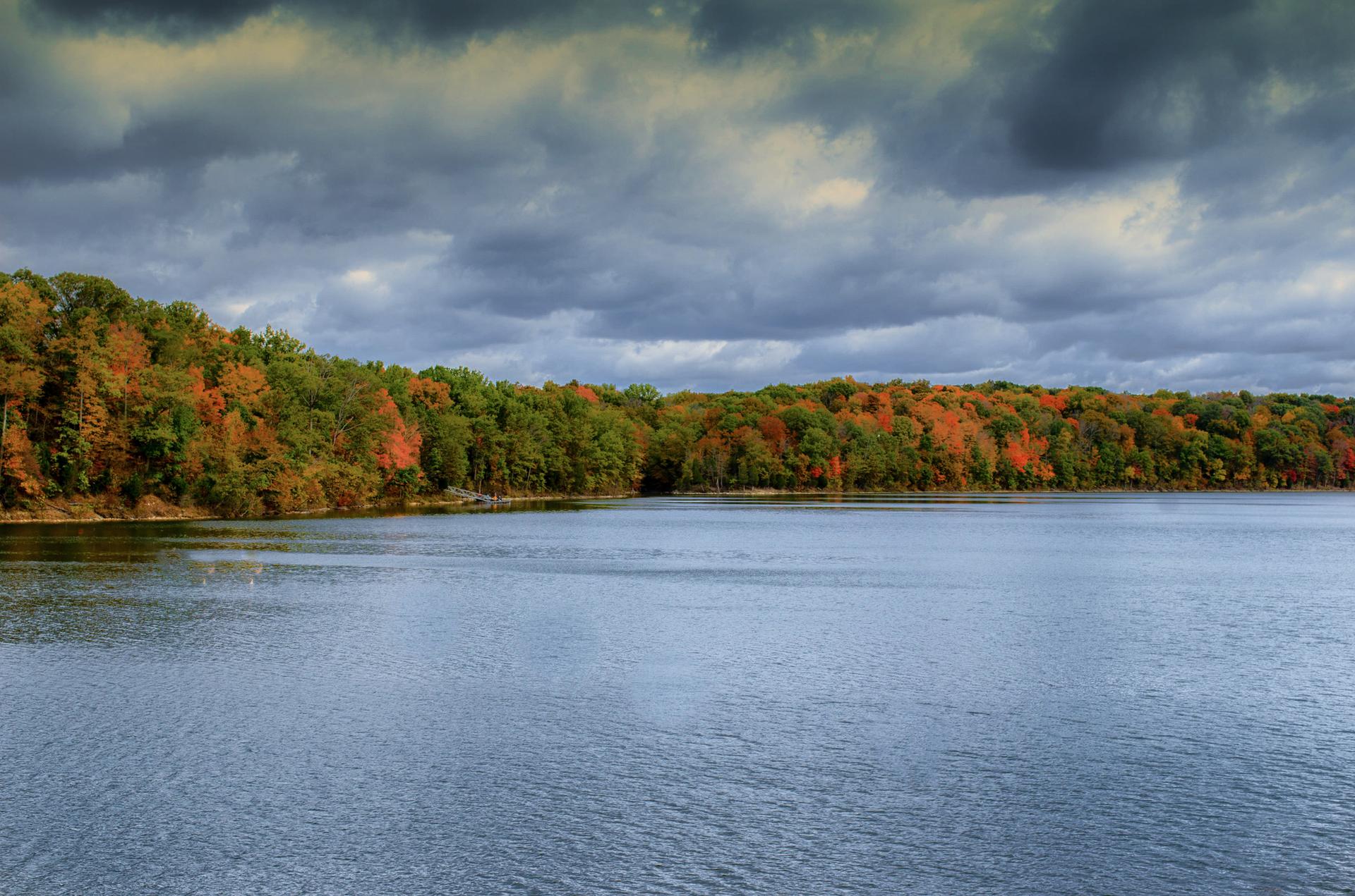 A lake with a colorful autumn forest under a dramatic cloudy sky.