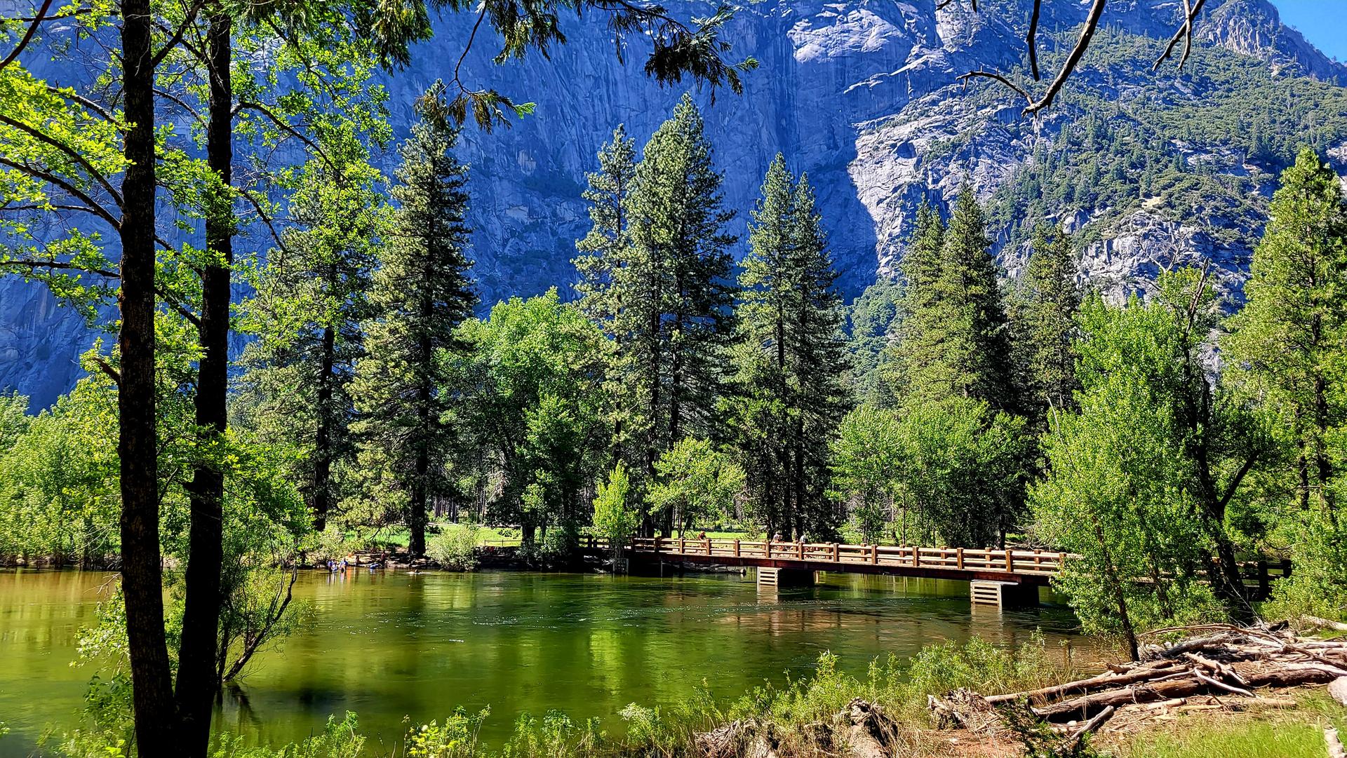 A wooden bridge spans a green river, surrounded by lush trees and mountains.