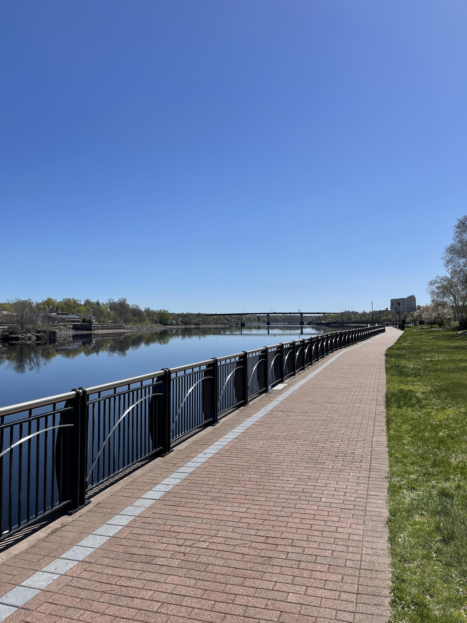 Paved riverwalk path with a black fence, calm river, bright sunny day.