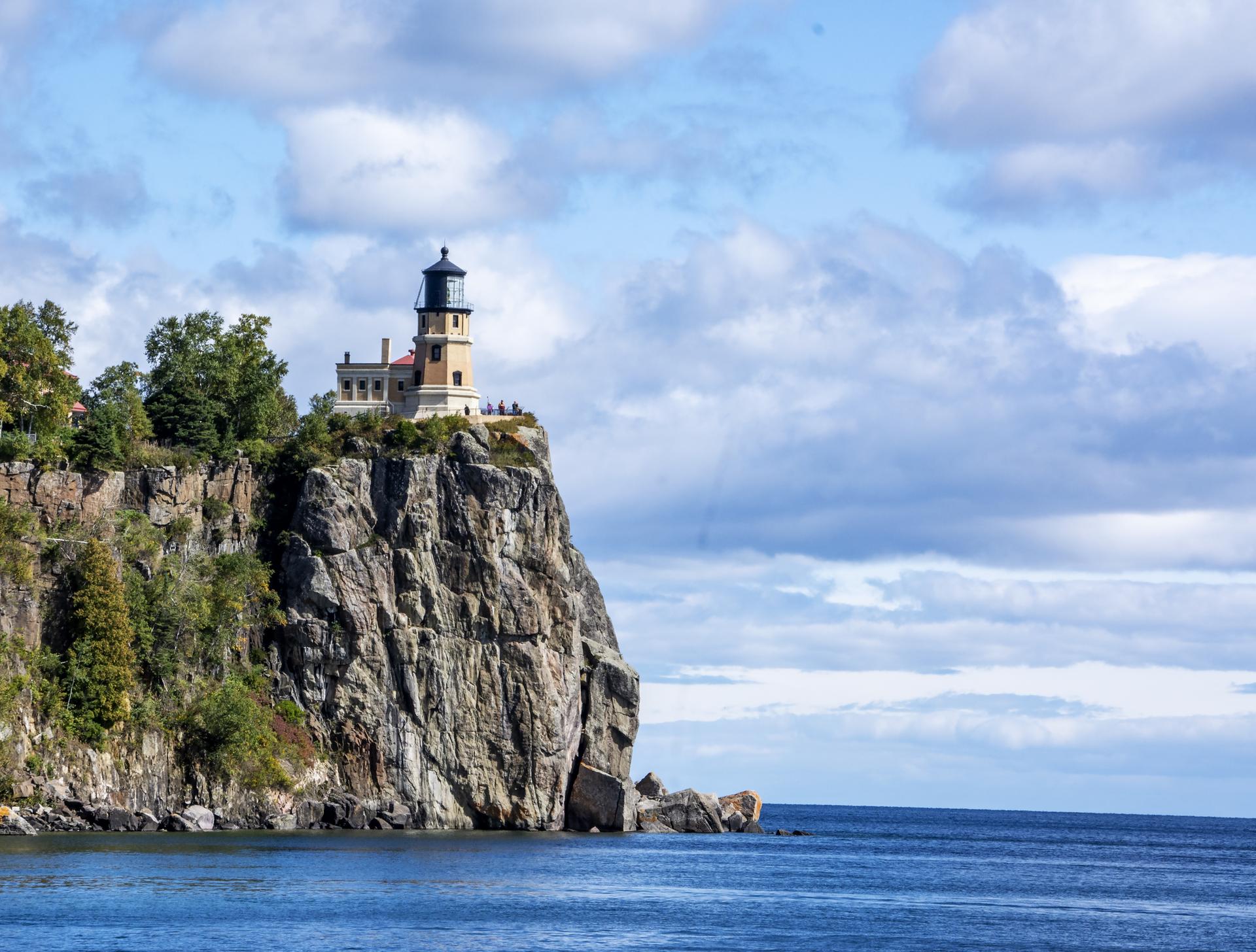 Lighthouse on a rocky cliff overlooking blue water under a cloudy sky.