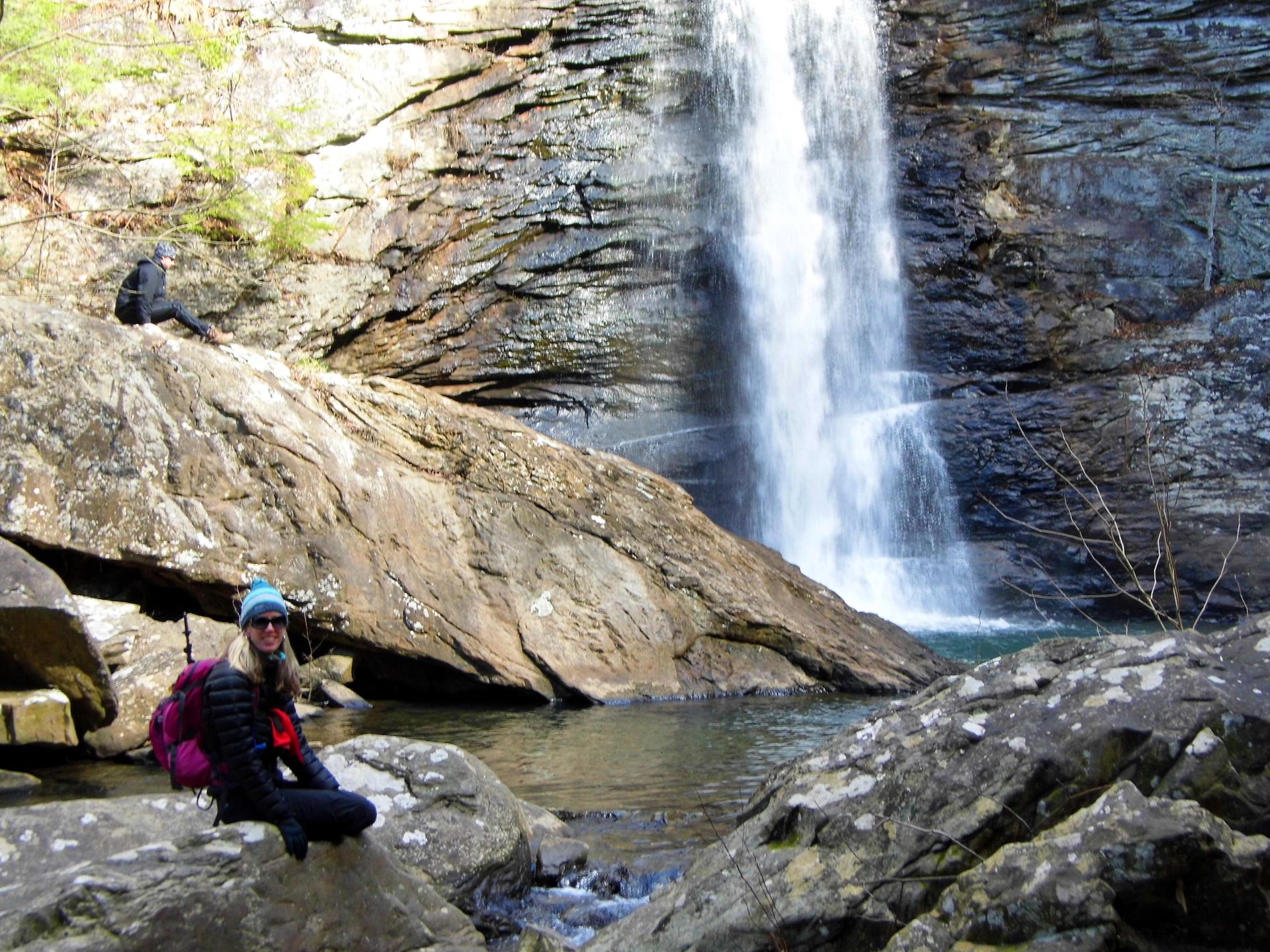 Two people near a powerful waterfall cascading into a rocky pool.