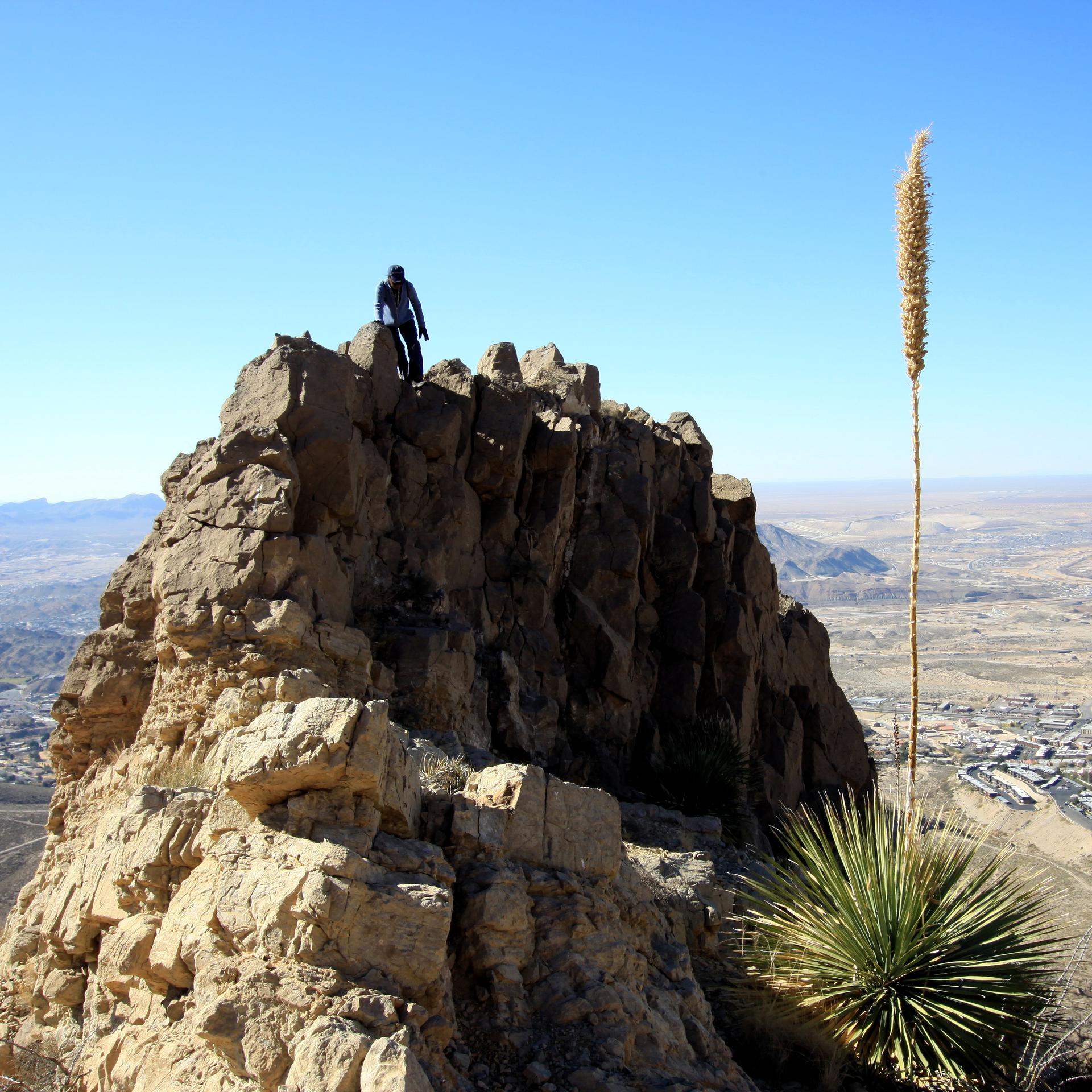 Person stands atop a rocky peak overlooking a vast desert and city.