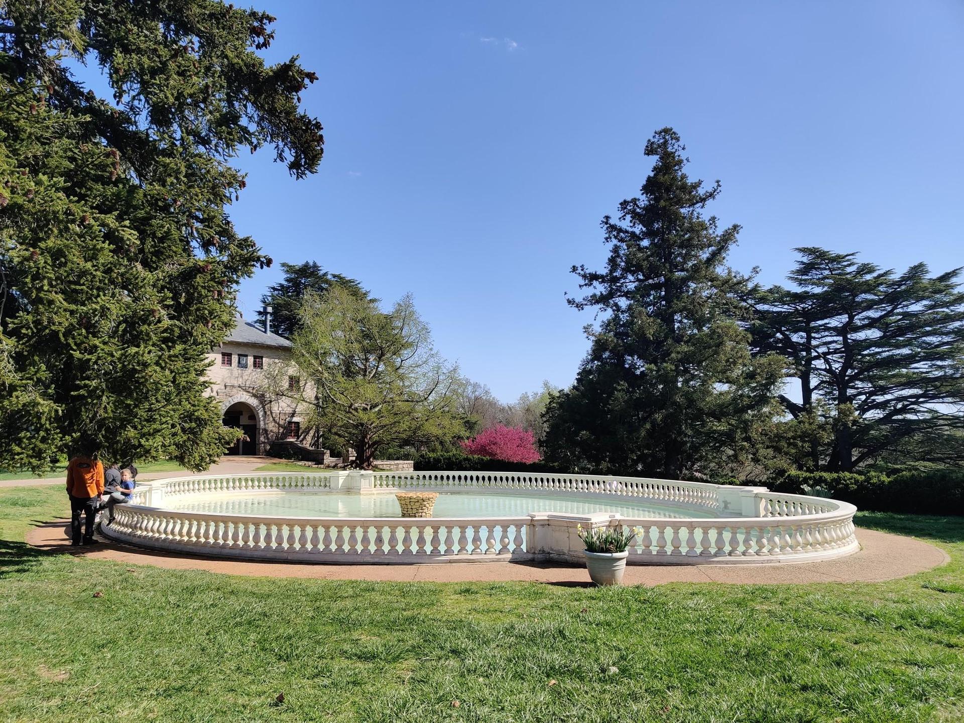 Sunny view of an oval fountain with a person and large trees.
