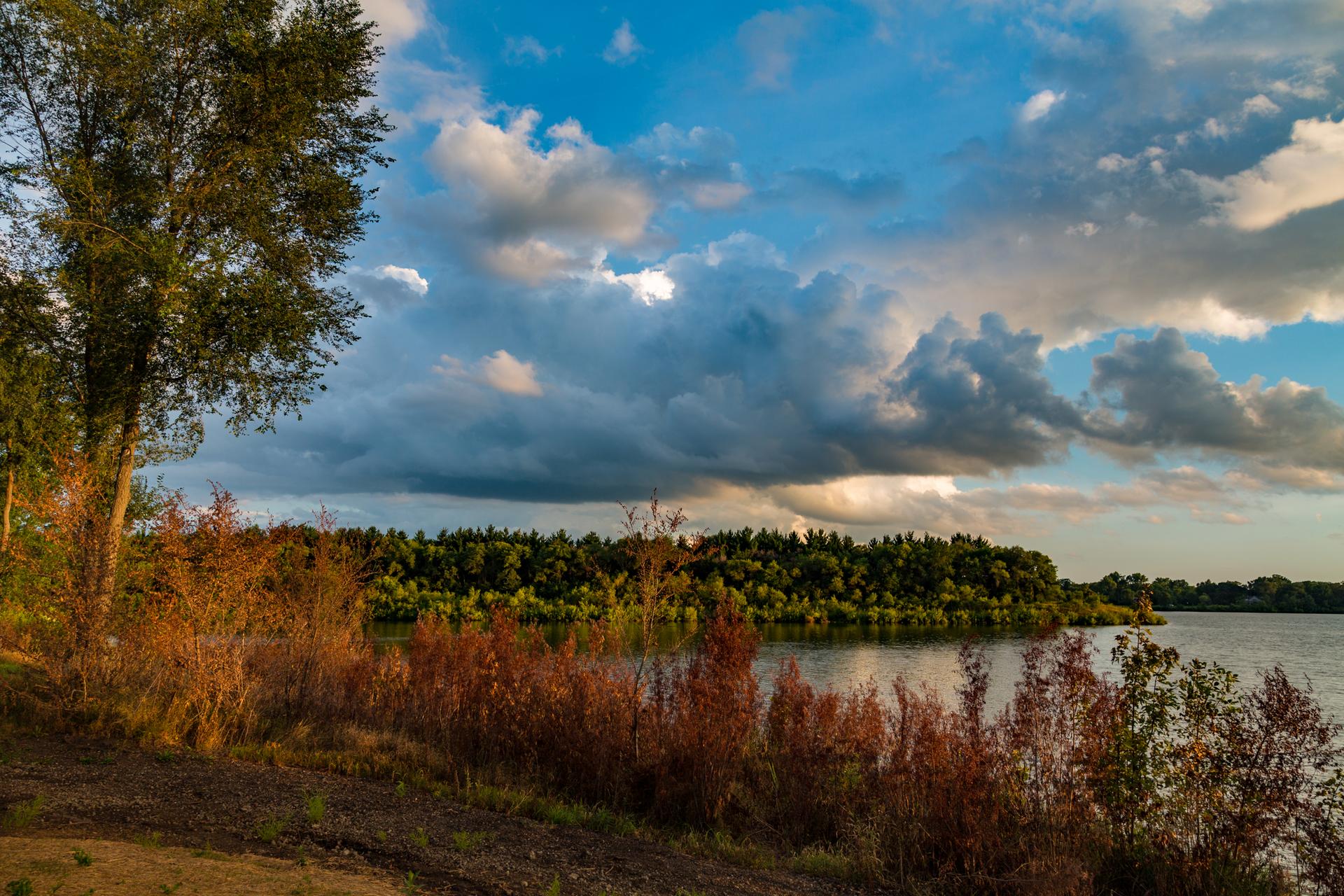 Lakeside view with warm light on autumn foliage and dramatic clouds overhead.
