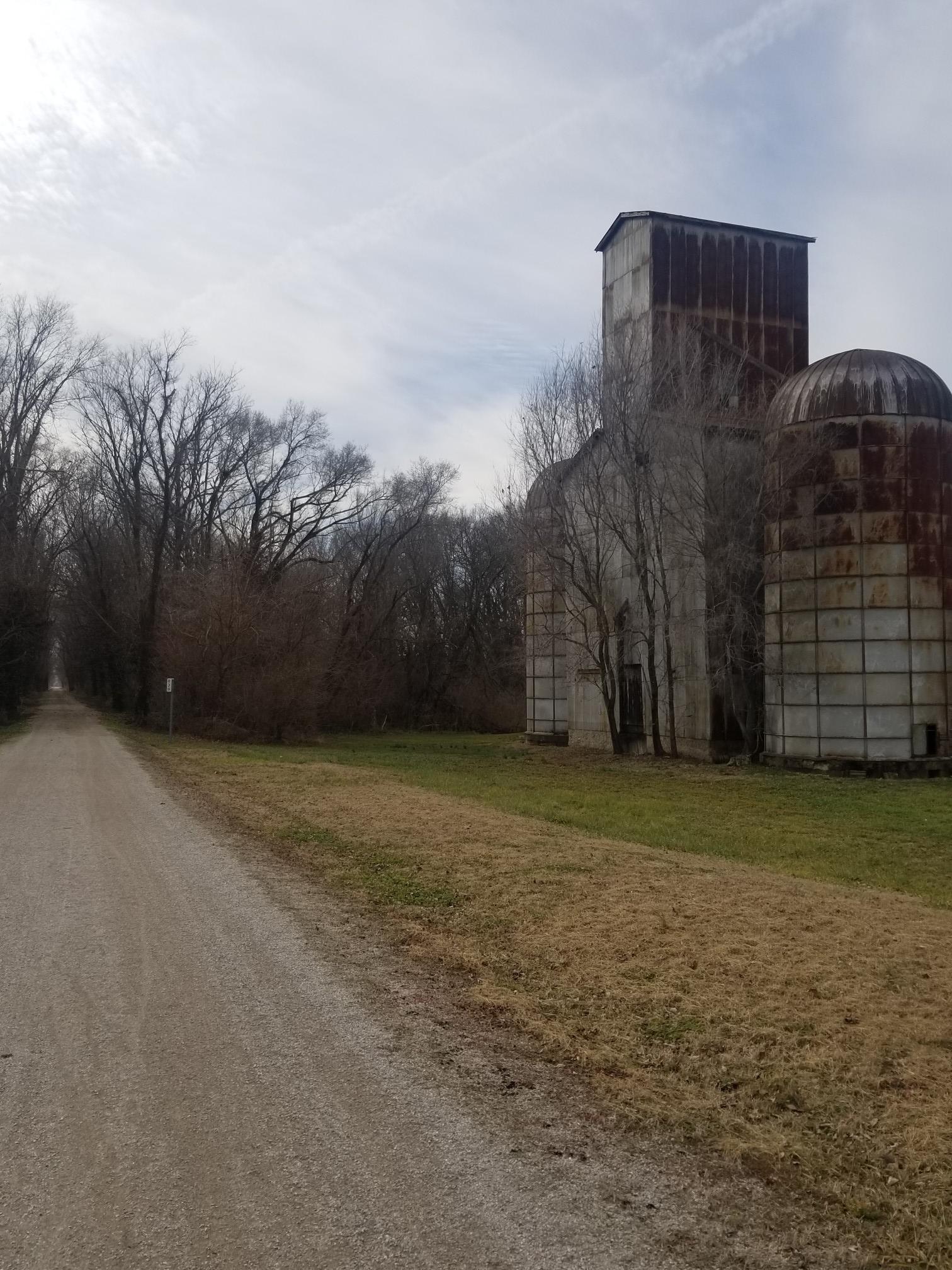 A dirt path runs alongside old, rusty industrial buildings and bare trees.