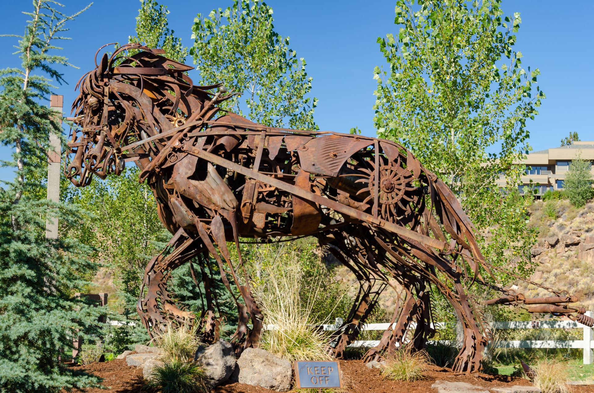 A large, intricate metal horse sculpture stands in a sunny garden.