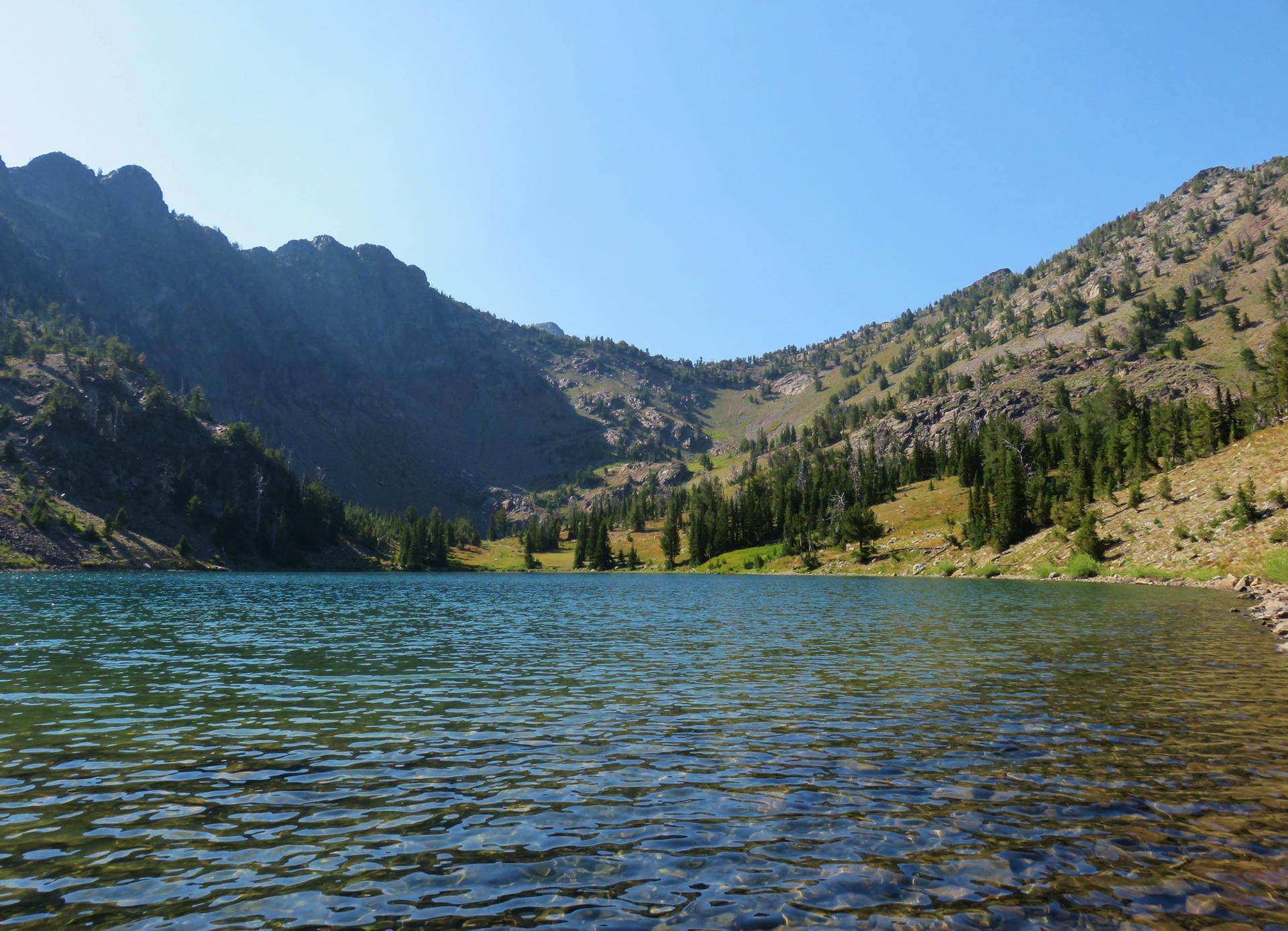 Clear mountain lake surrounded by steep, rocky, forested slopes under a blue sky.