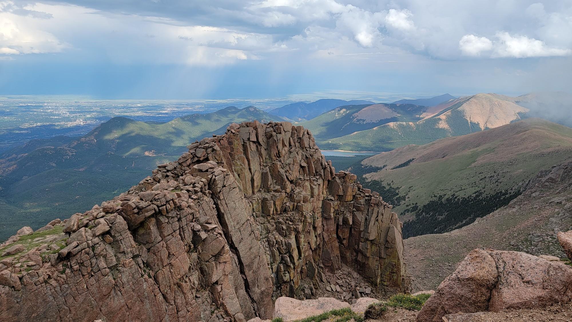 Rugged rocky peak overlooks green mountains and a distant valley under cloudy skies.