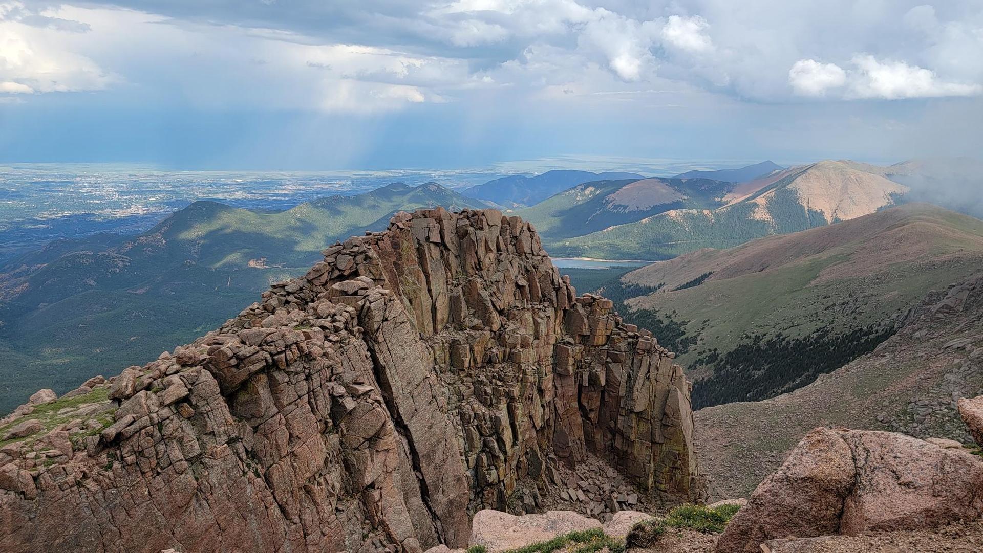 Rugged rocky peak overlooks green mountains and a distant valley under cloudy skies.