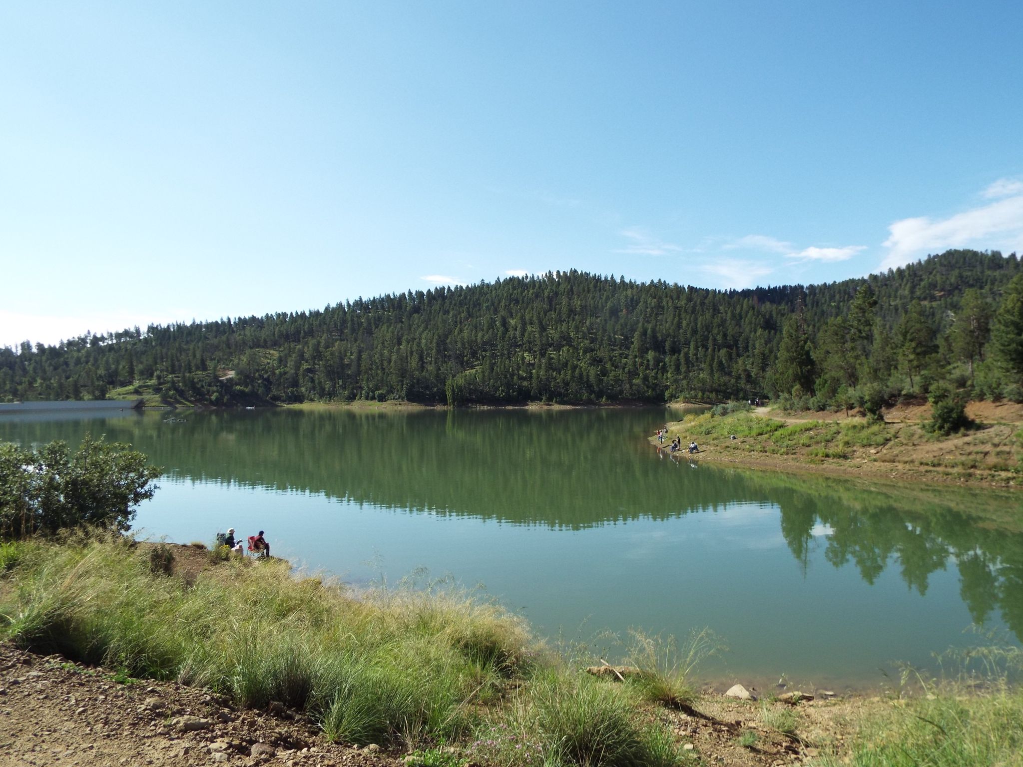 Two people sit by a calm lake surrounded by green forested hills. - Grindstone Lake loop from Ruidoso
