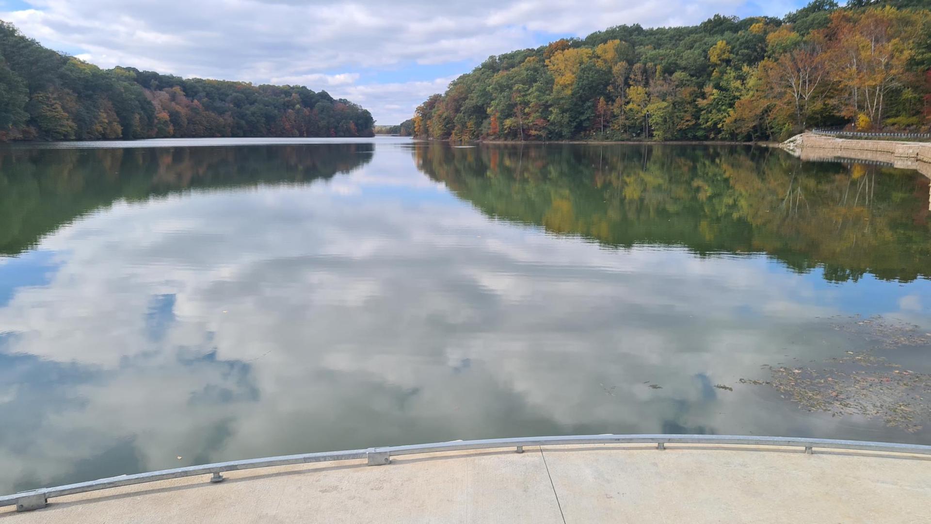 A serene lake reflecting cloudy skies, flanked by hills with autumn foliage, from a concrete platform.