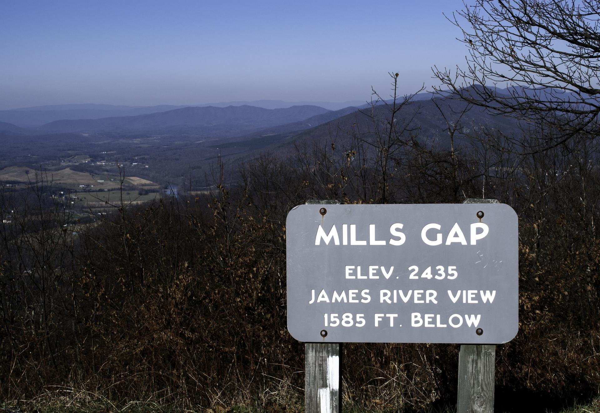 A gray sign in front of a wide, hazy mountain landscape on a clear day.