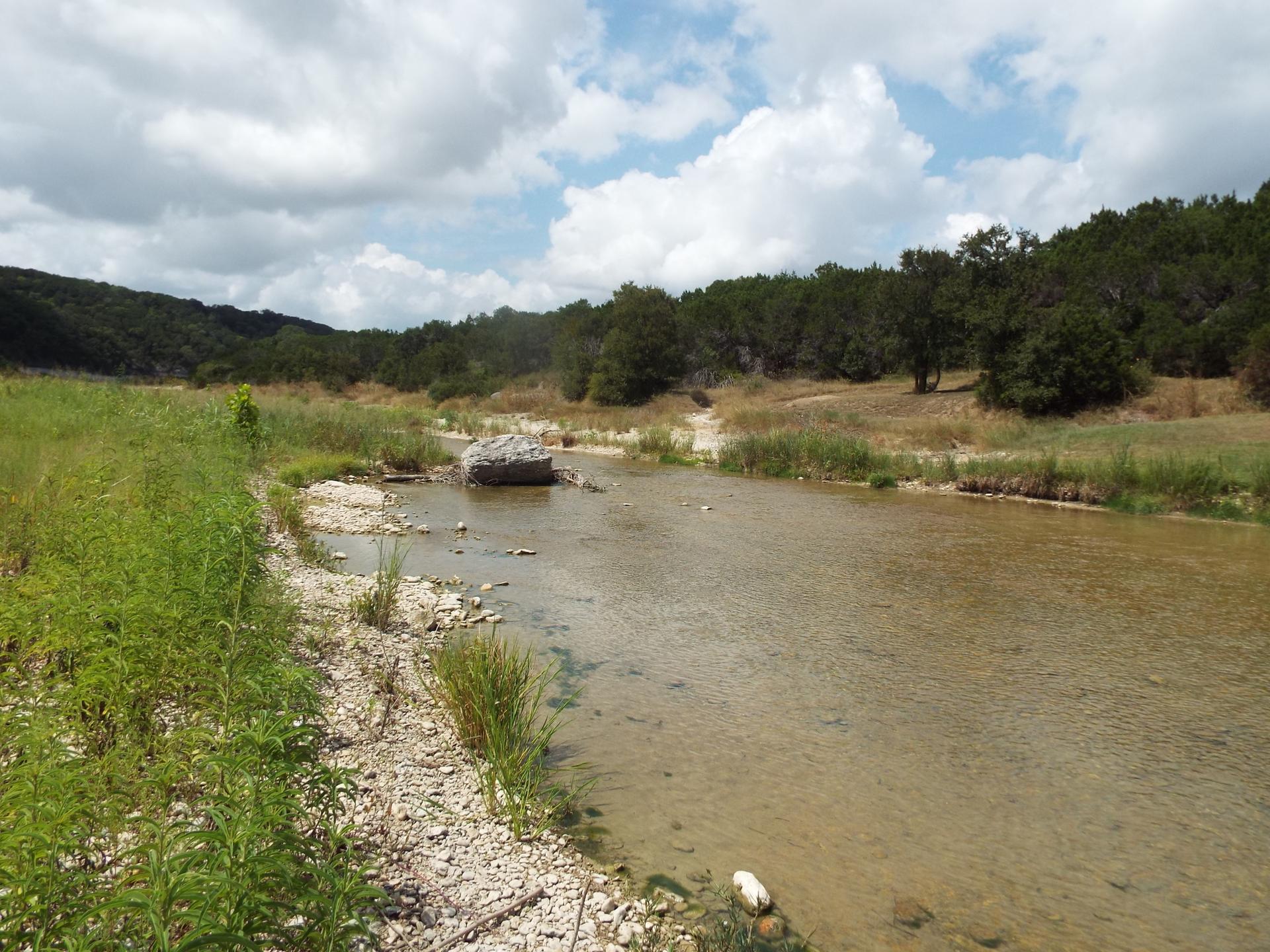 Shallow river with rocky bed, green banks, and wooded hills under a cloudy sky.