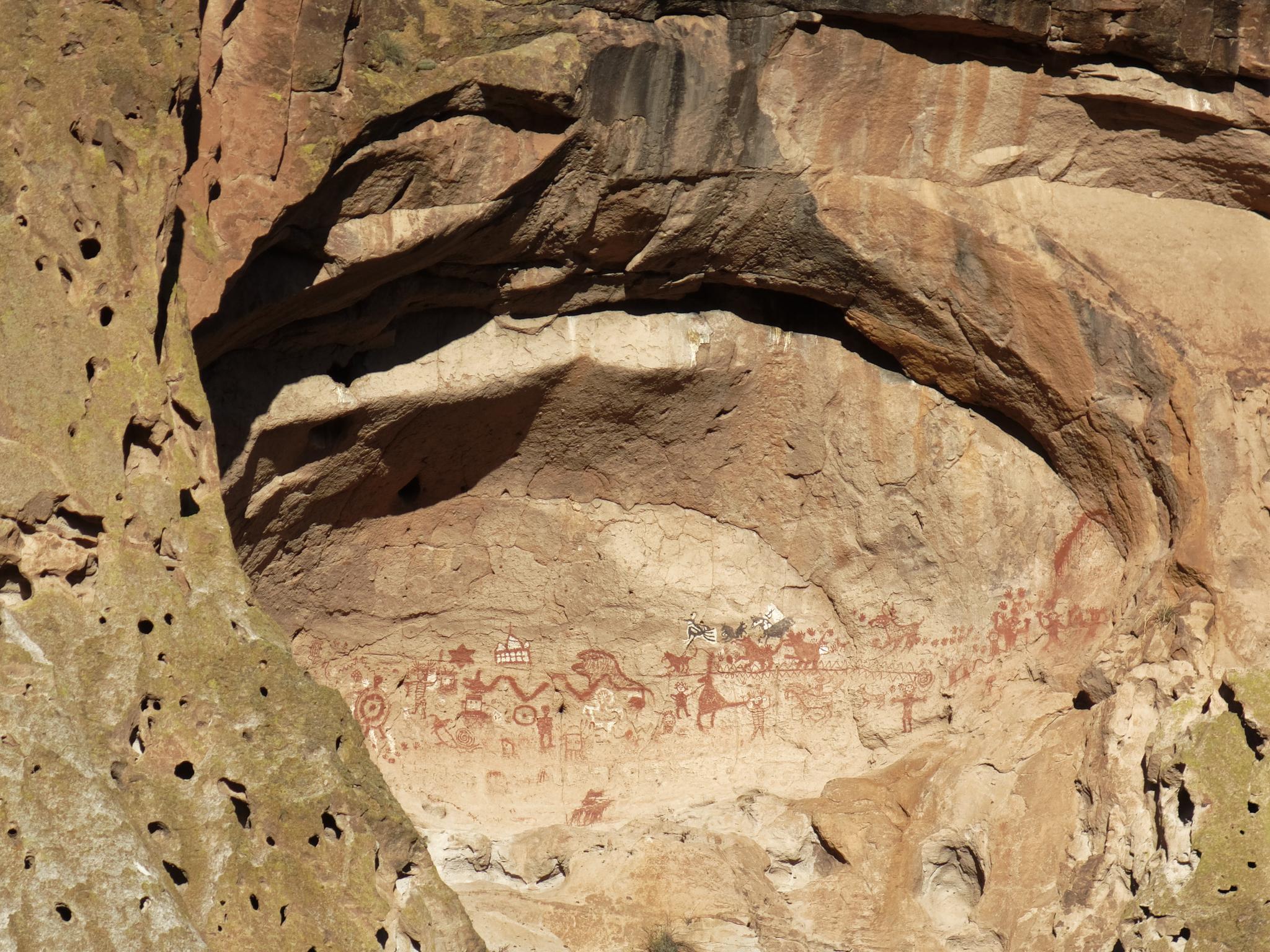 Ancient red pictographs adorn a large rock overhang on a sunny cliff face.