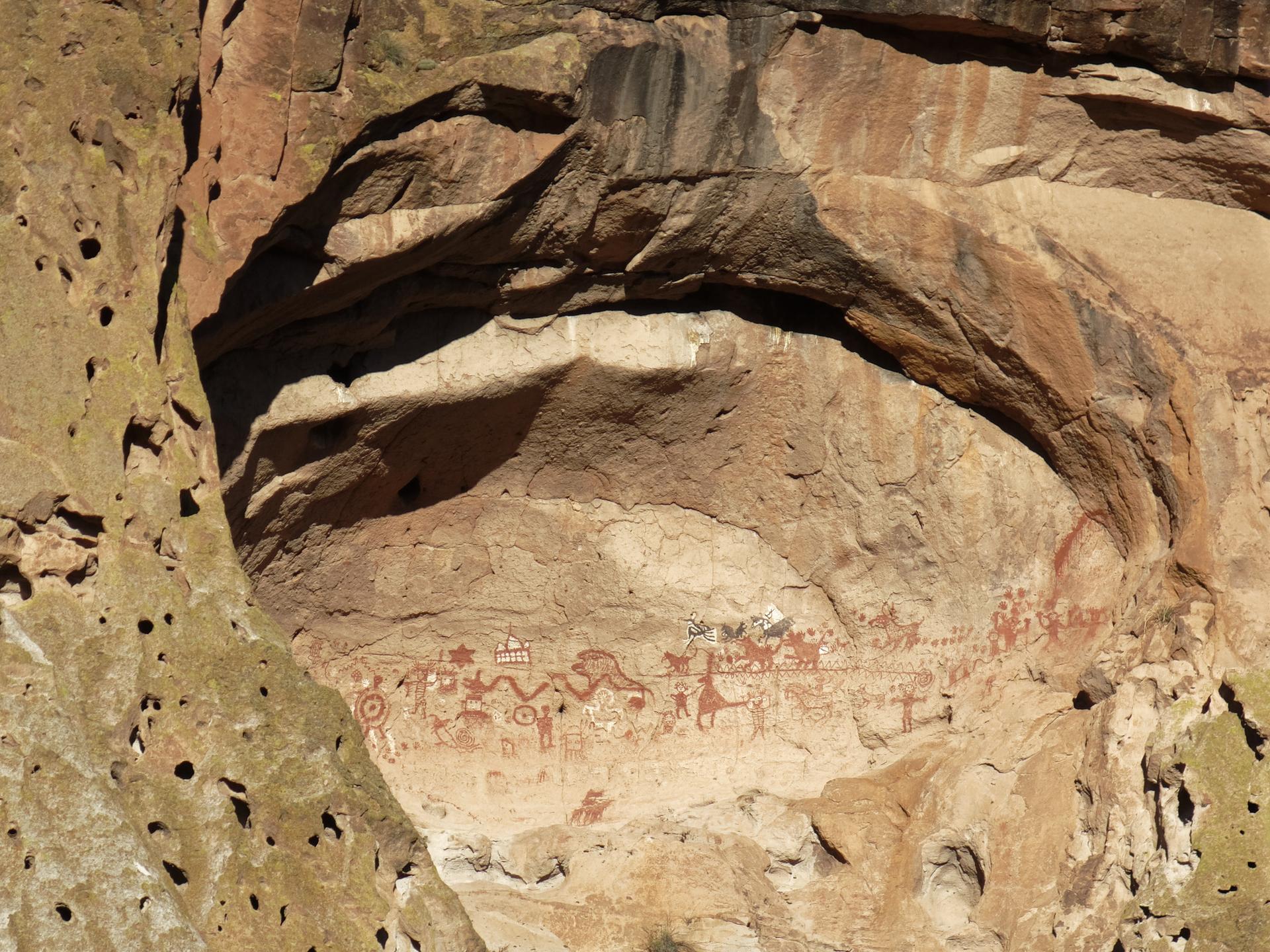 Ancient red pictographs adorn a large rock overhang on a sunny cliff face.