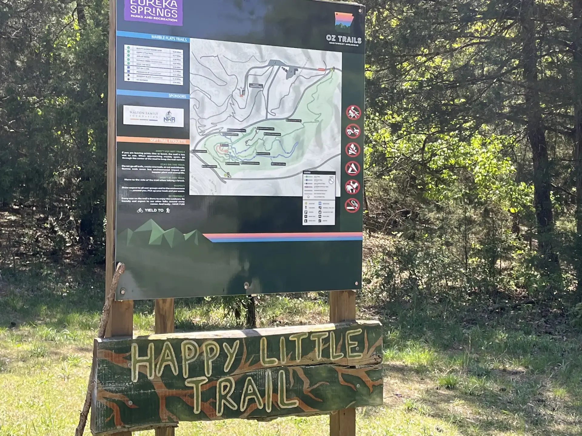 A large trail map sign stands at the edge of a sunny forest.