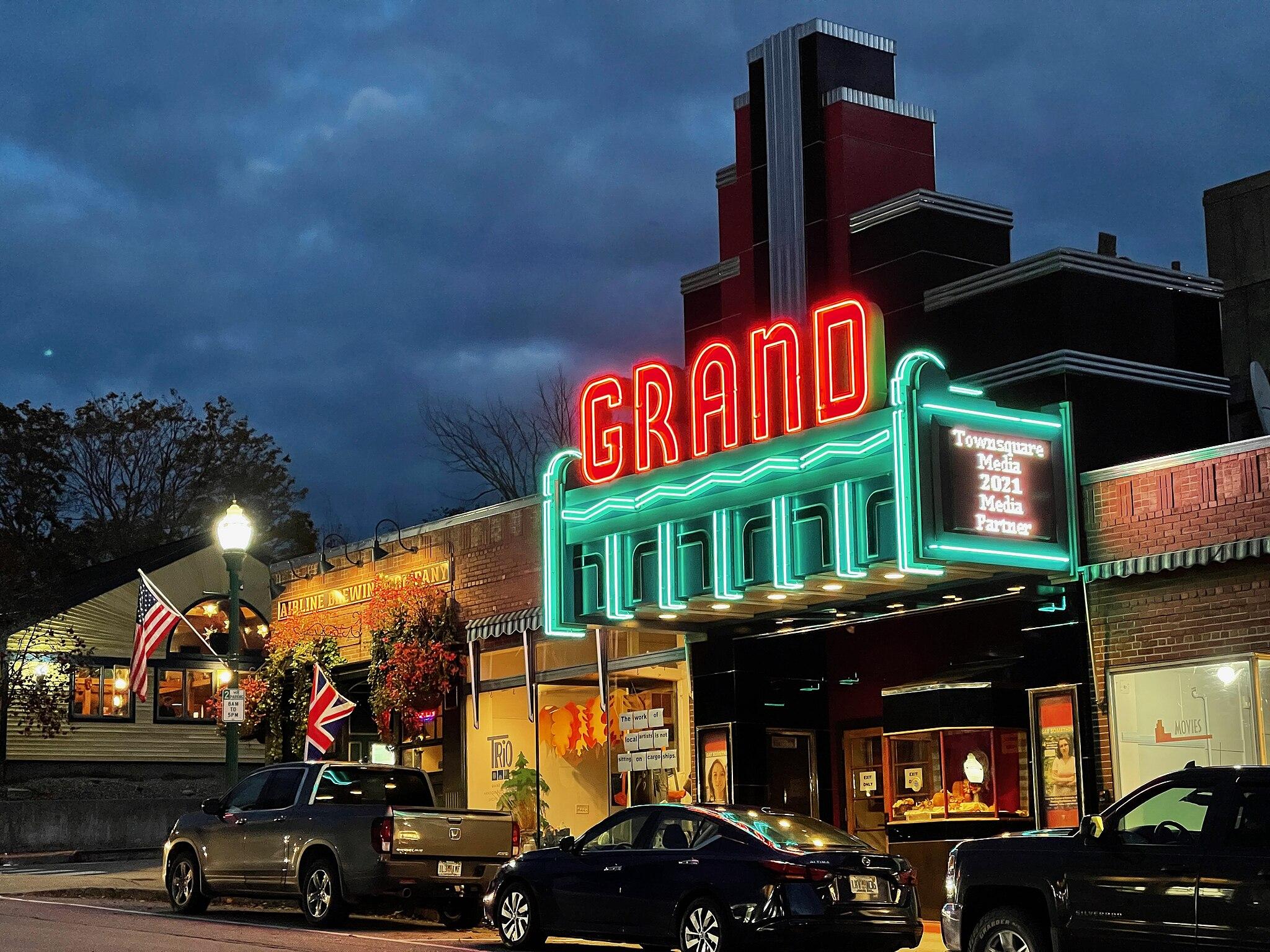 Neon-lit 'GRAND' theater marquee at dusk with cars parked on the street.