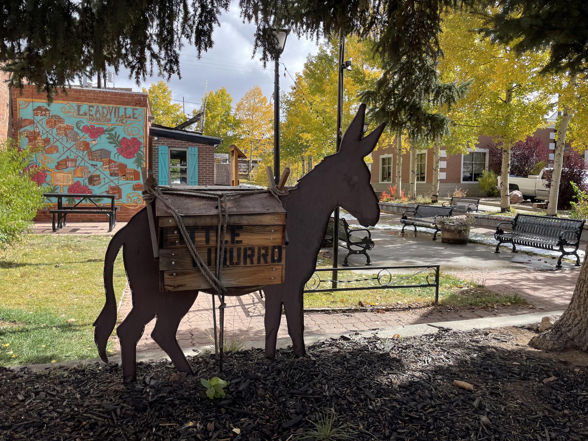 Metal donkey sculpture with pack in a park with autumn trees.