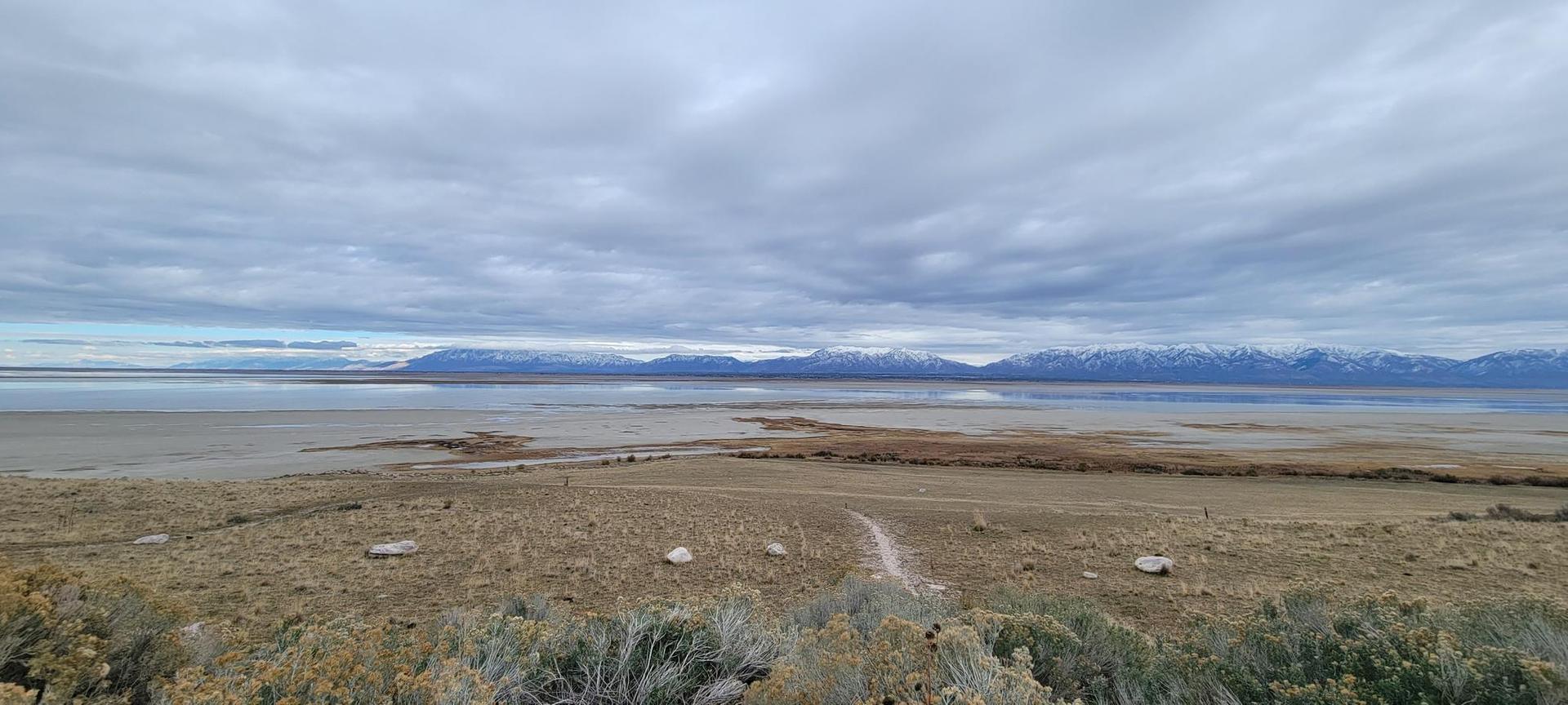 Overcast view of a vast lake, distant snowy mountains, and a winding dirt path.