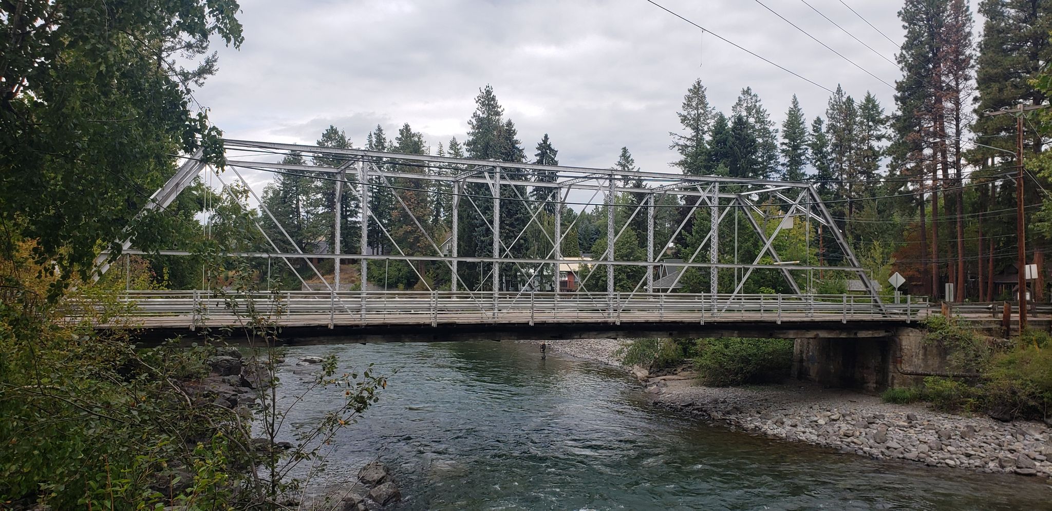 A metal truss bridge spans a river with forested banks under a cloudy sky. - Swan River Bridge – Swan River Trail loop from Echo Lake