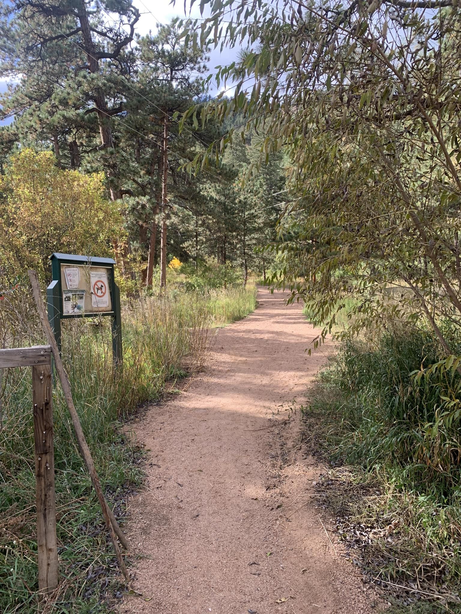 A dirt path next to a trail sign, surrounded by tall grass and trees. - Palmer Reservoir Trailhead – Palmer Lake loop from Palmer Lake