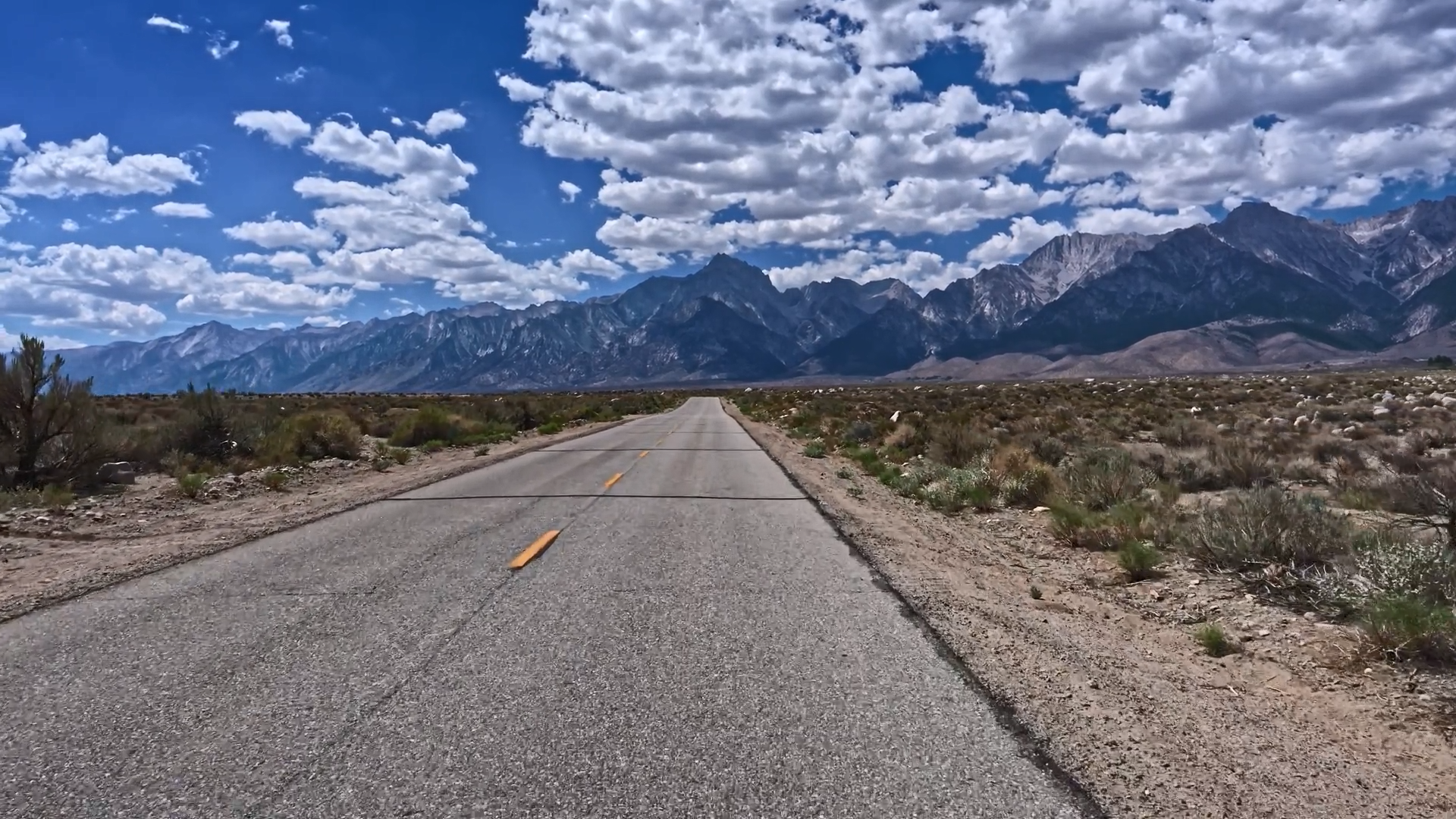 A long road stretches towards rugged mountains under a bright, cloudy sky.