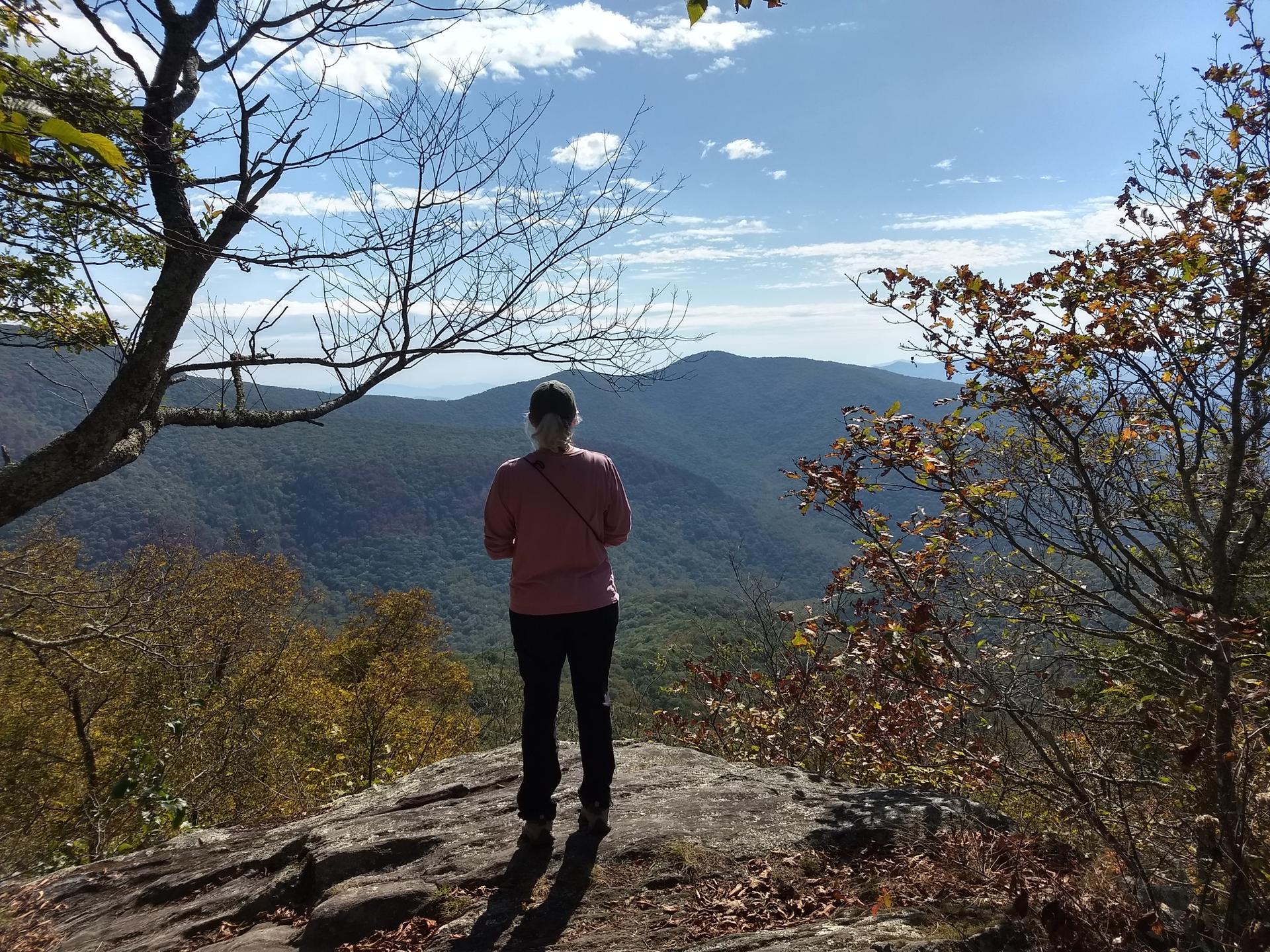 Person stands on rocky outcrop, admiring a vast mountain landscape.