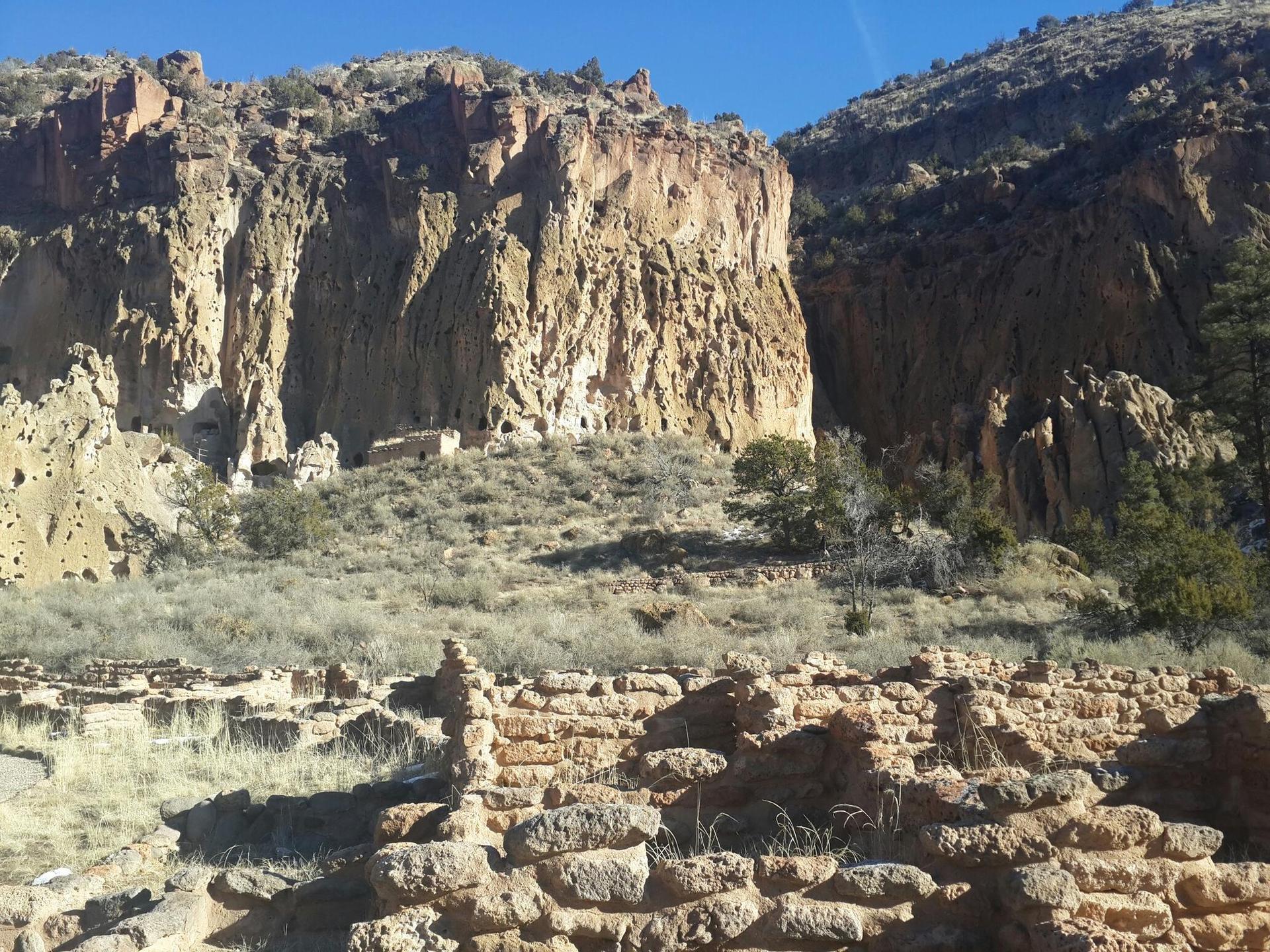 Ancient stone ruins in foreground, towering rocky cliffs with caves in background.