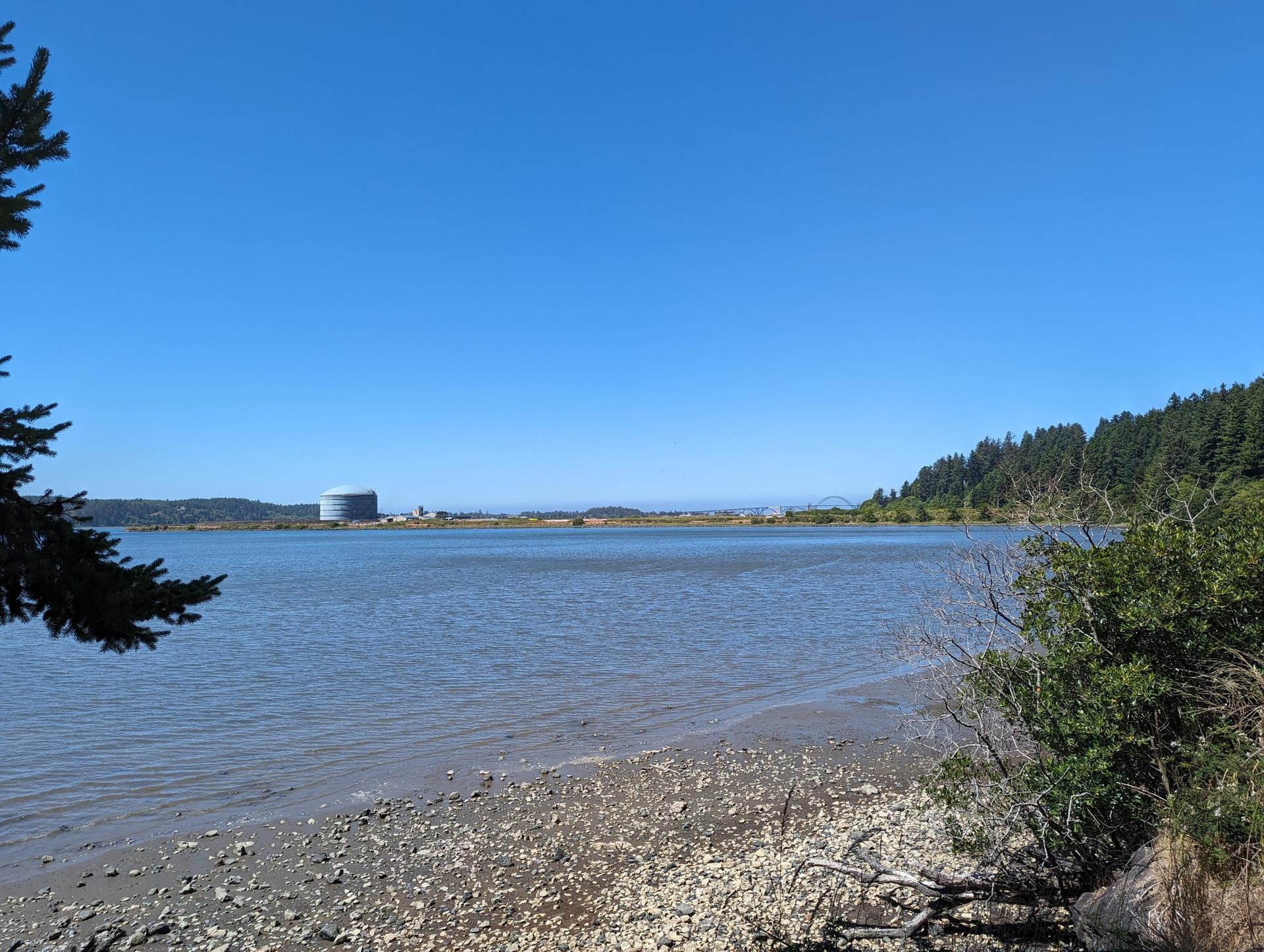 Calm water with a rocky shore, distant building, and forested hills.