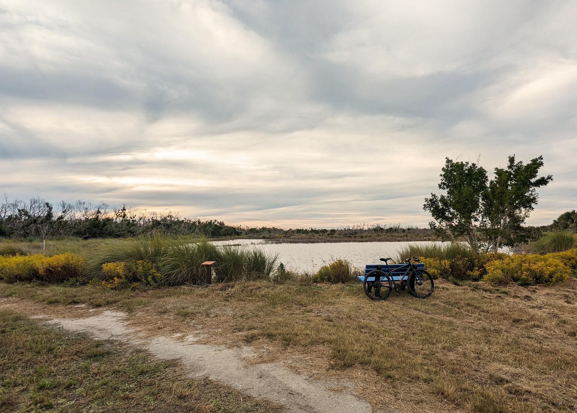 A bicycle with a cart sits by a calm lake under an overcast sky.