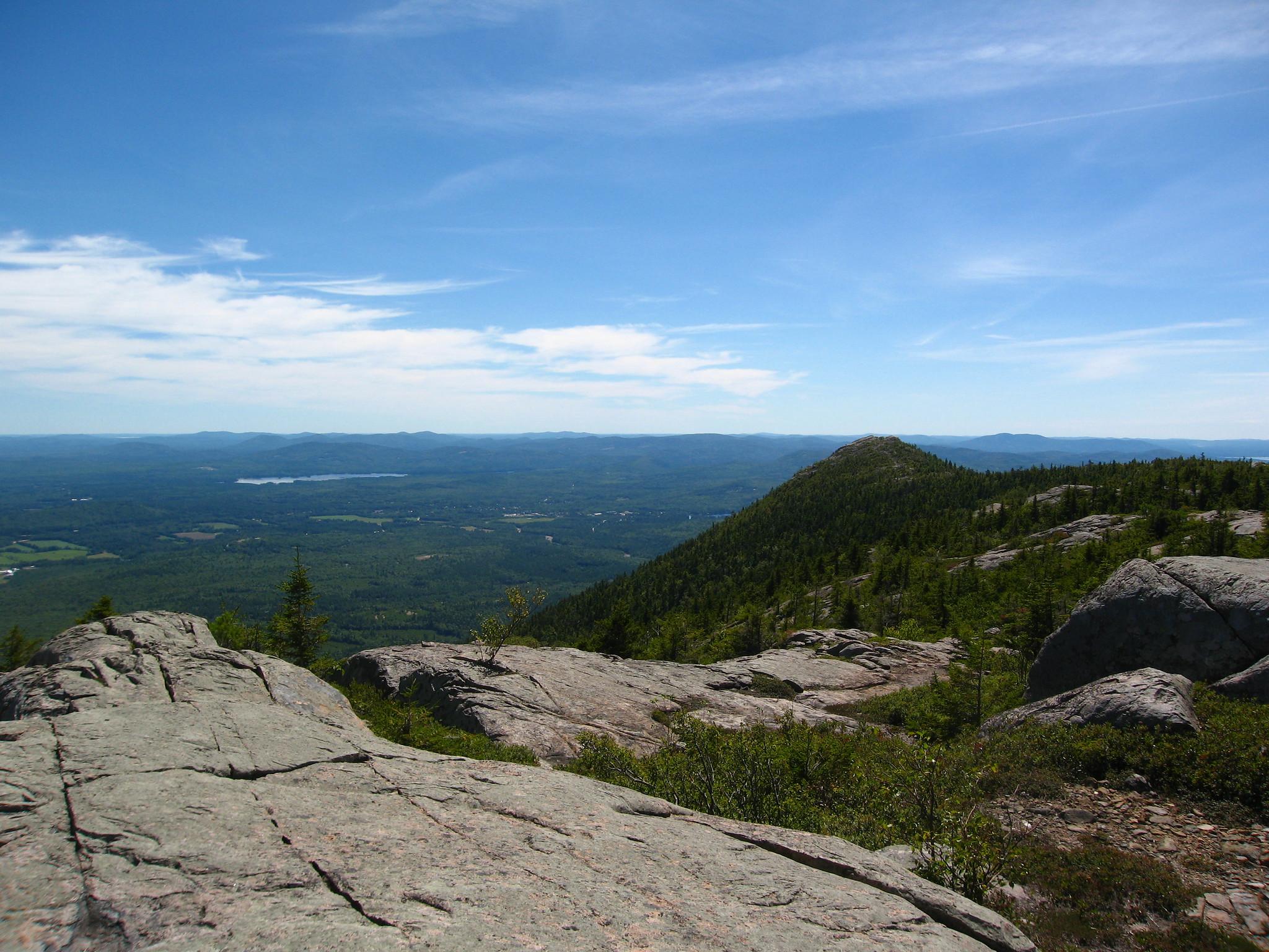 Sunny mountain vista with rocky foreground and distant forested peaks.