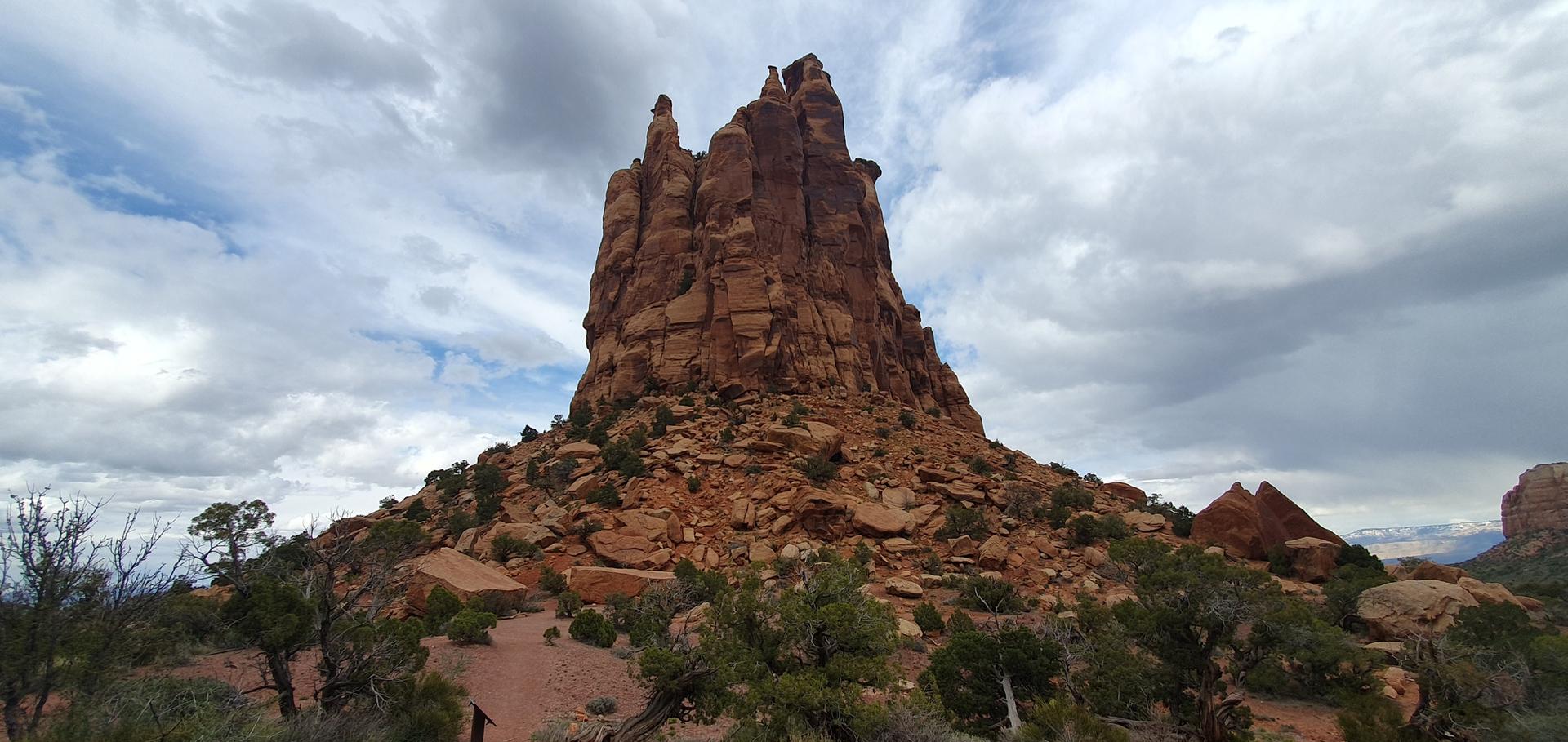A tall, rugged red rock spire dominates a desert landscape under a cloudy sky.