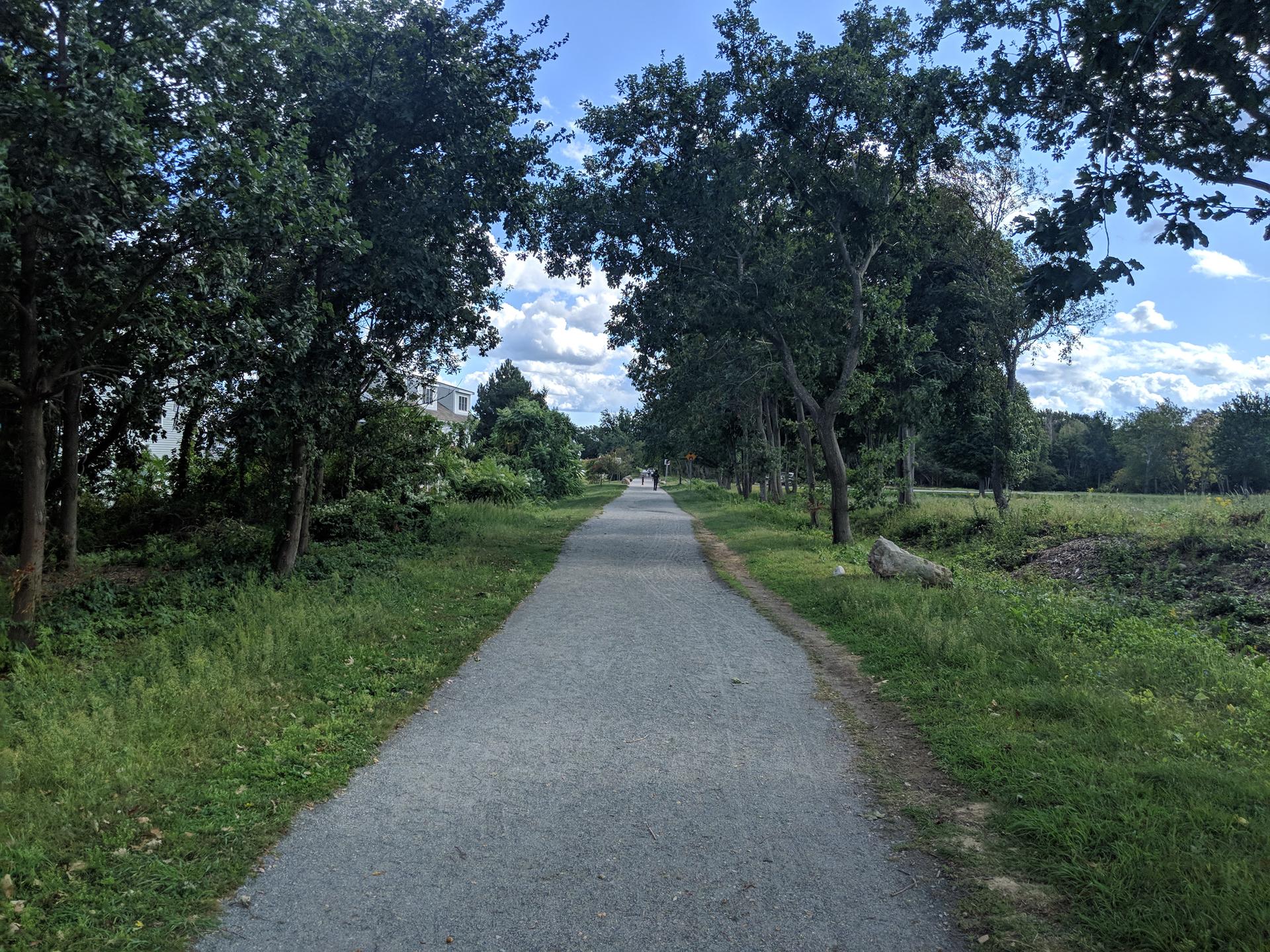 A paved path through green landscape under a bright sky, person walking.