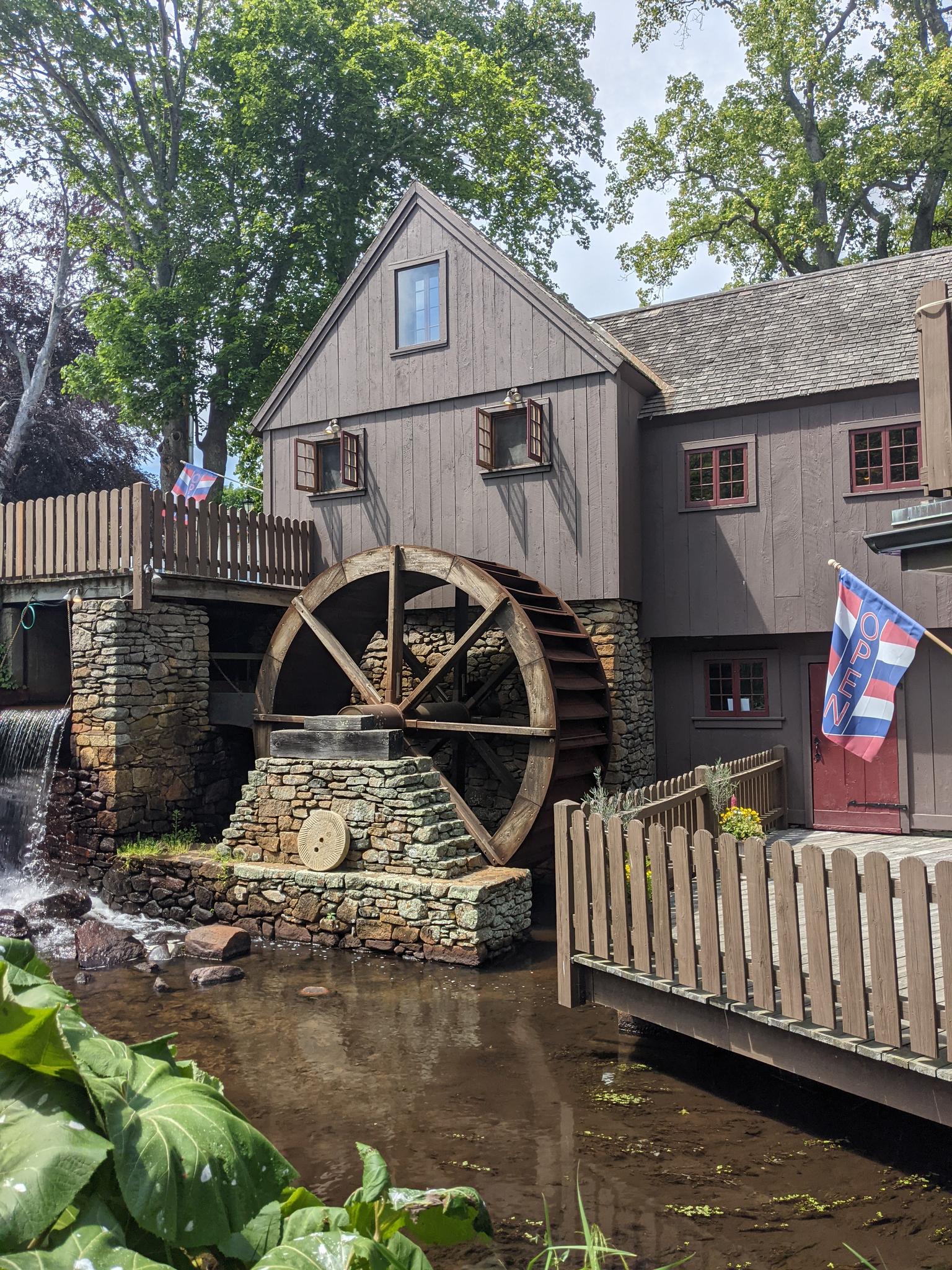 Large wooden water mill with a turning wheel, stone foundation, lush greenery.