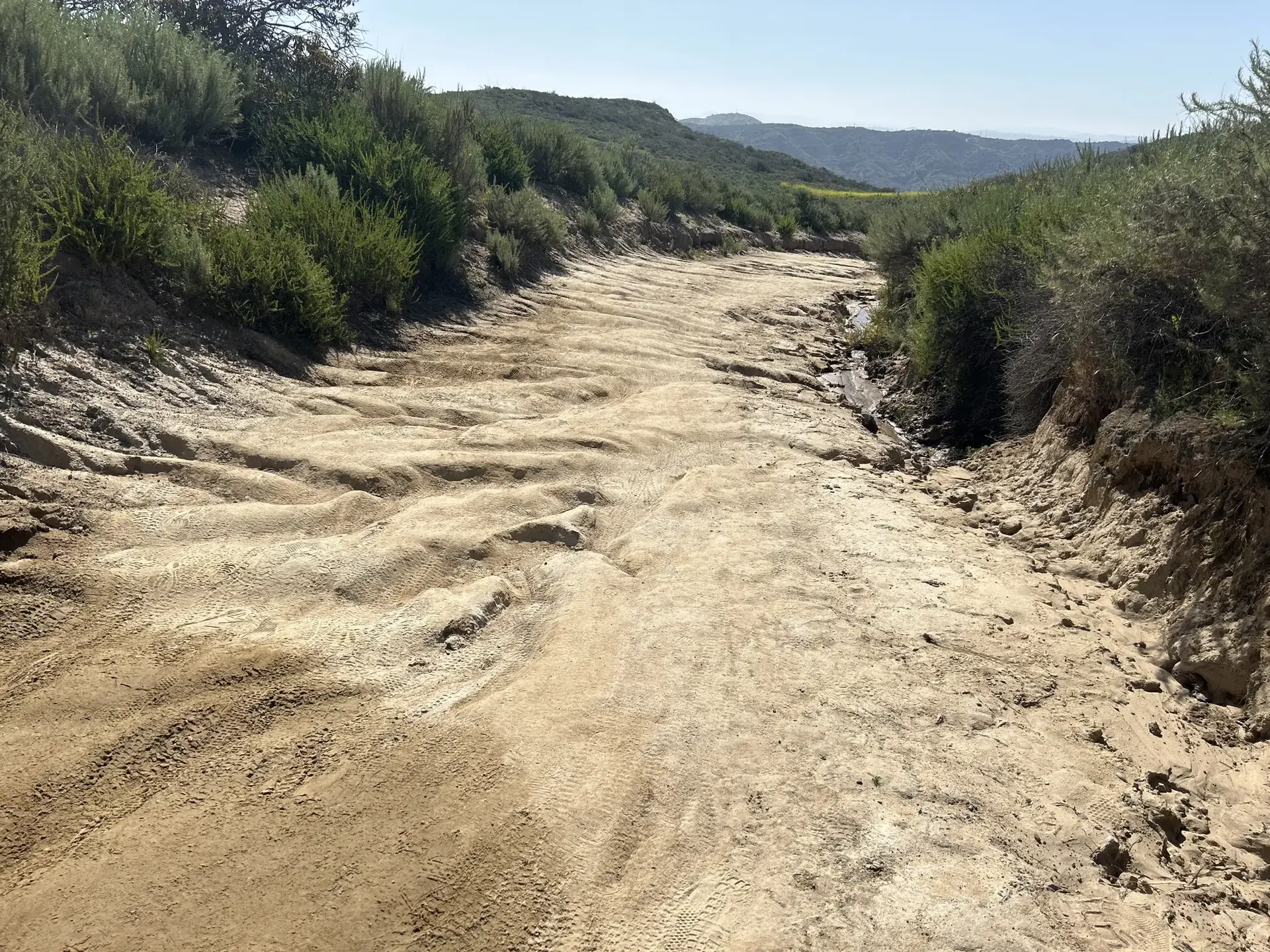 Wide, rutted dirt road through a brushy, hilly landscape.