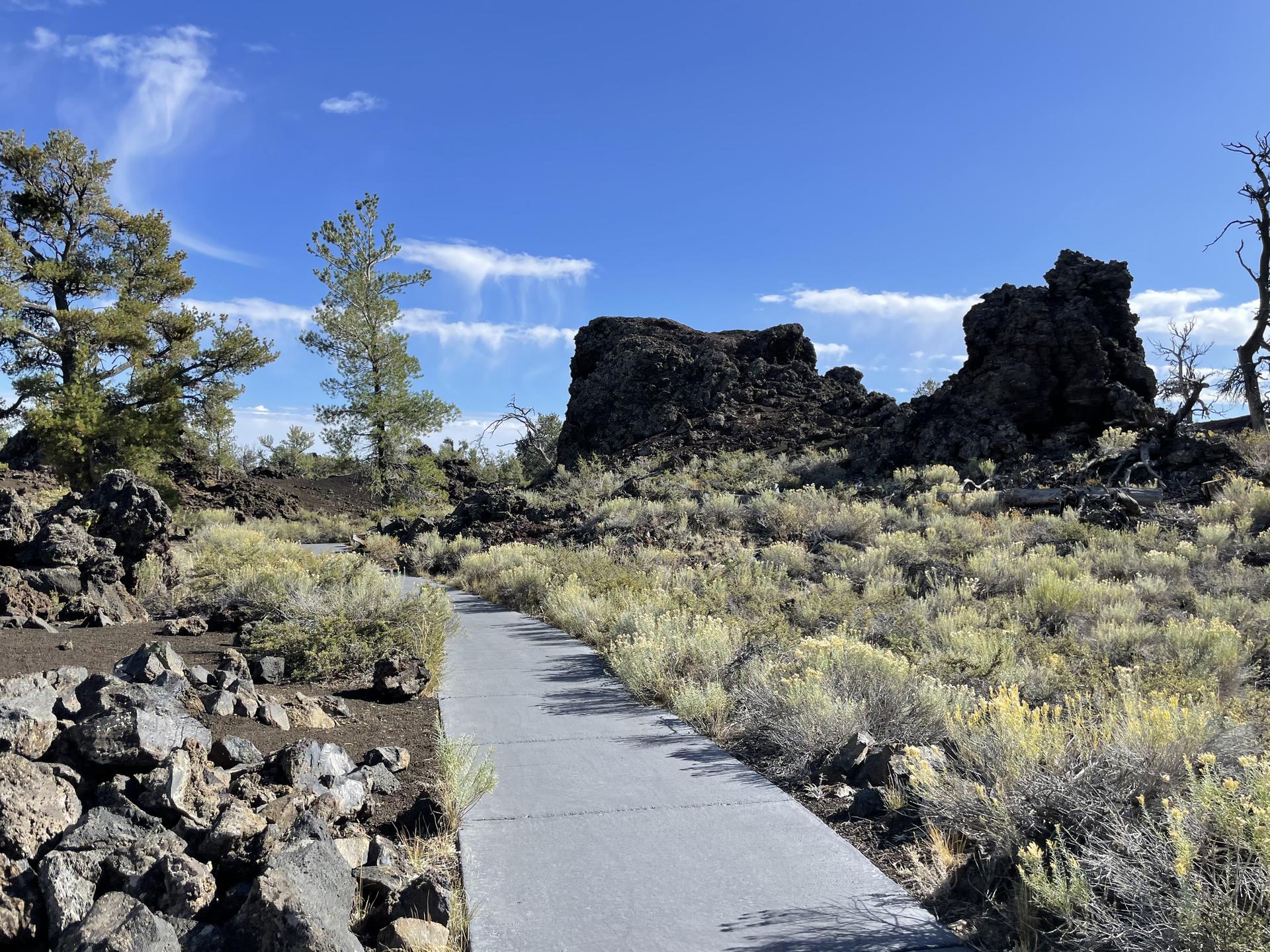 Paved path leads through rocky terrain with shrubs and large dark formations.