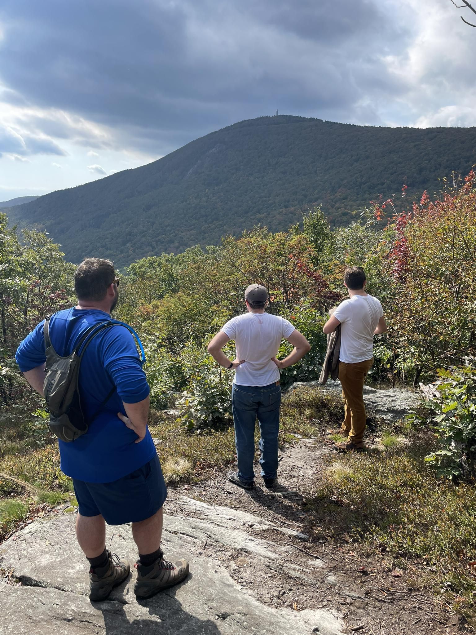 Three men stand on a rocky overlook, admiring a vast forested mountain range. - Bellows Pipe Trail to Ragged Mountain