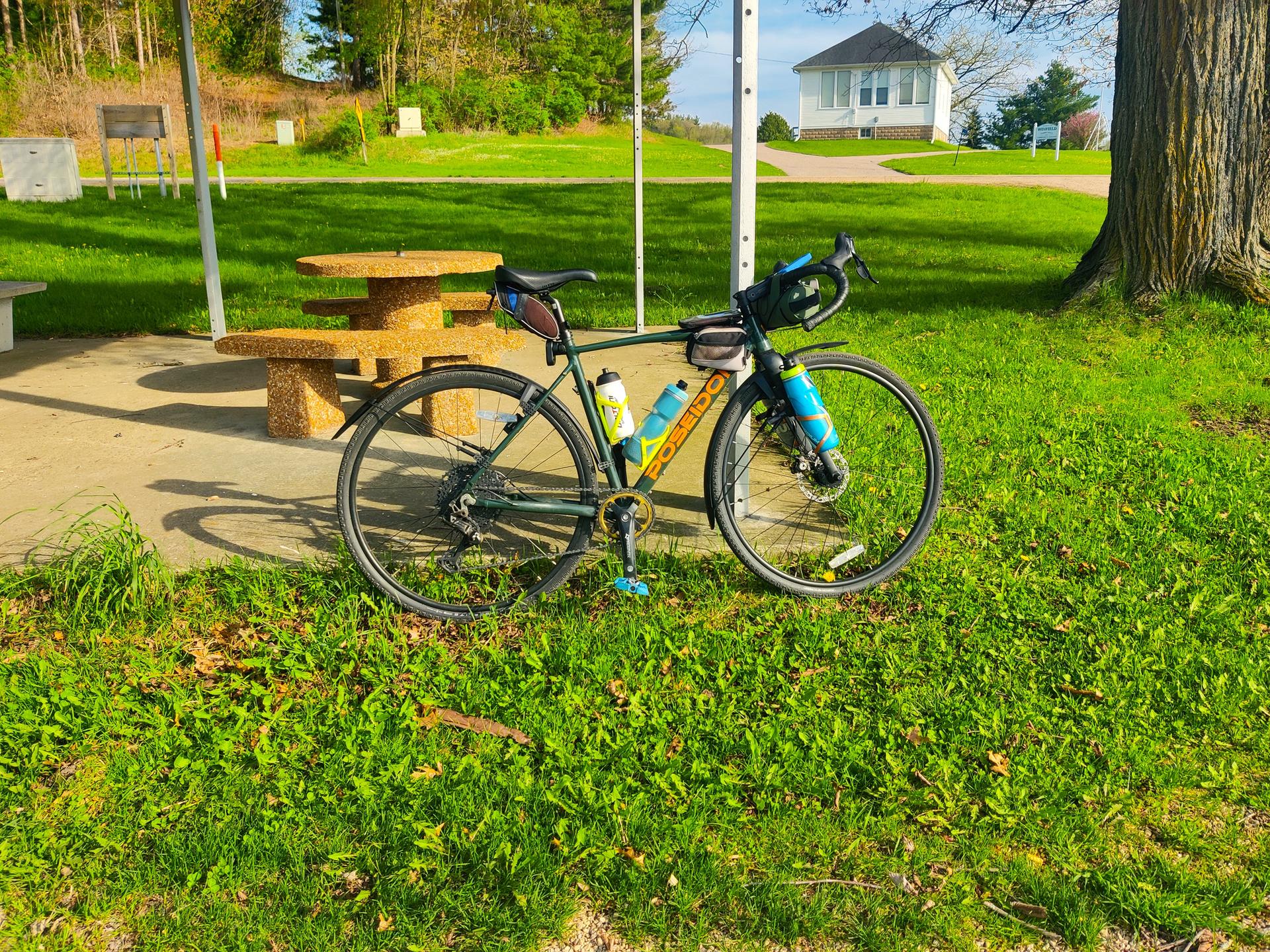 A bicycle with bags parked on grass next to a picnic table on a sunny day.