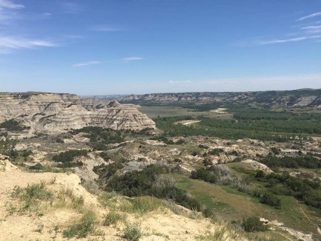 Sunny, expansive badlands with layered rock formations, green trees, and a winding river.