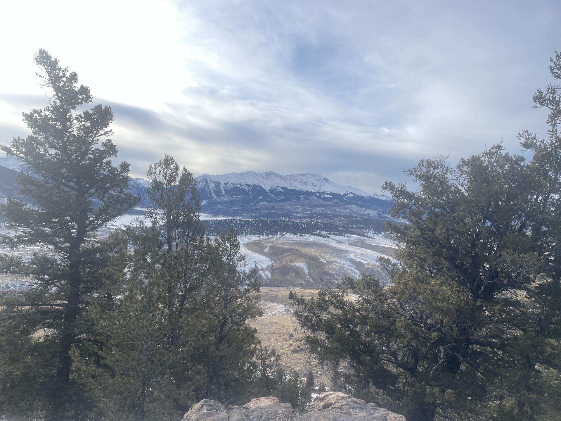 Evergreen trees frame a panoramic view of a snowy valley and distant mountains.