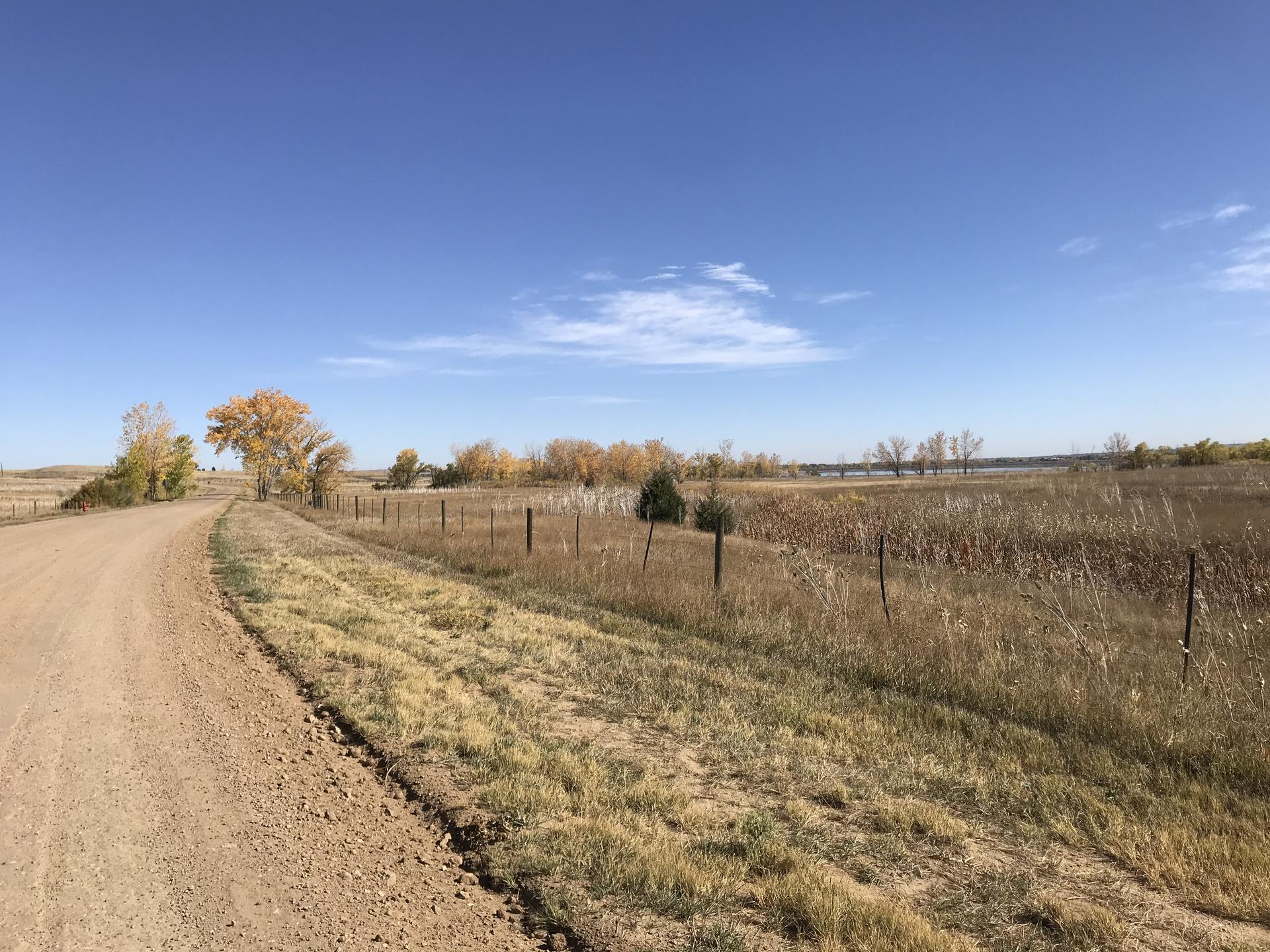 Sunny rural landscape with a dirt road, dry grass, and colorful autumn trees.